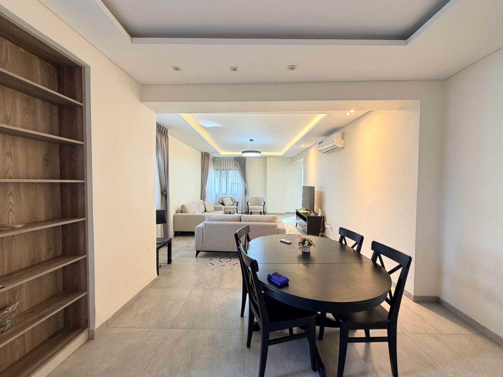 173a018b-b225-4b72-8668-18478bce0c96 Open-plan living and dining area with a round black dining table in the foreground, beige sofas, and large window with curtains in the background.