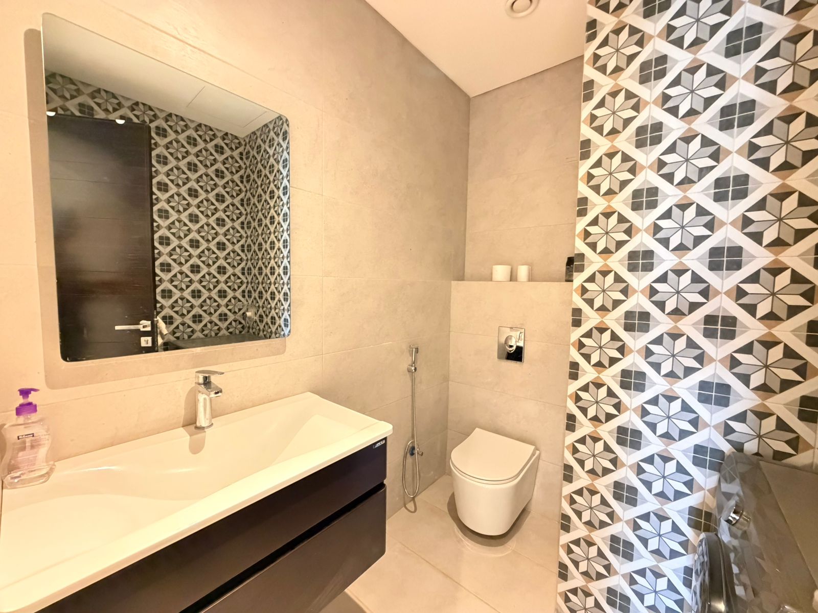 1cb51e9d-d826-4867-b067-7a4c248e8813 Contemporary bathroom with geometric patterned tile wall, large mirror above a white sink and a wall-hung toilet with handheld bidet spray.