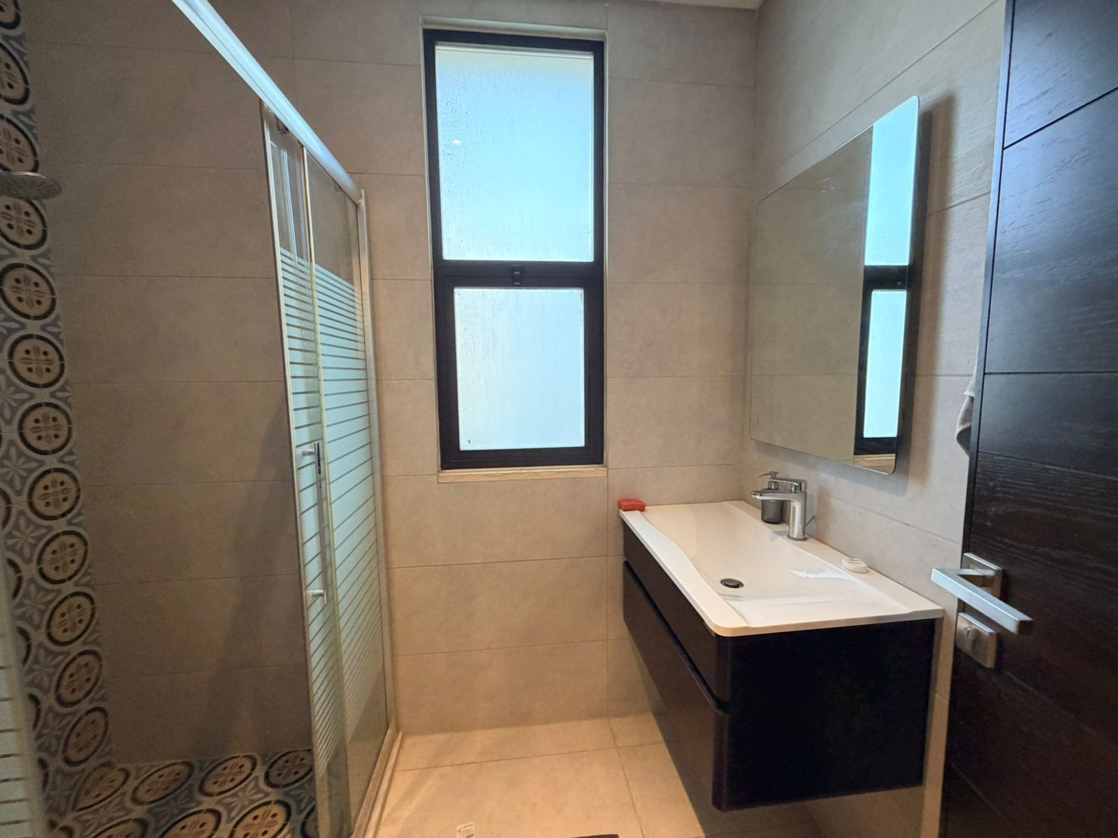 1f5348eb-033d-42b7-8f09-7323f4f88251 Bathroom with glass shower enclosure on left, frosted window in center, and white sink with dark vanity beneath a large mirror on the right.