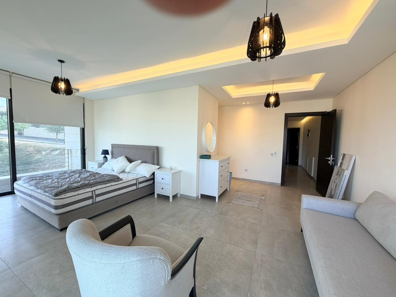 260b87f4-a5ce-44c3-b1bb-a24507f28f5e Spacious modern bedroom with a queen bed, two white nightstands, and a dresser with a round mirror against a pale wall.