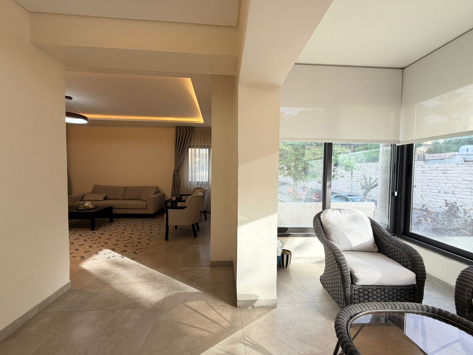 606d4e94-063c-407a-859f-0c2ec06a17df Open living area with a beige sofa, armchairs, and dark coffee table atop a patterned rug; large windows bring in sunlight and overlook a sunlit balcony with wicker seating.