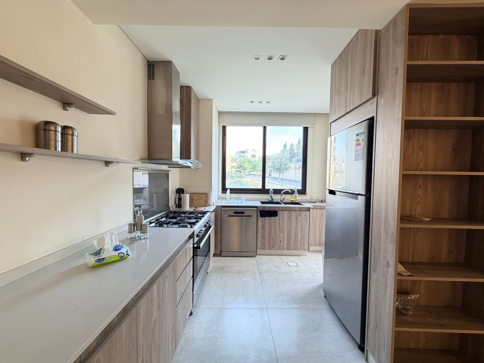 9f286a67-319b-4071-9ed6-a26028b9a27d Modern kitchen with light wood cabinets, stainless steel appliances, and a large window overlooking trees outside.