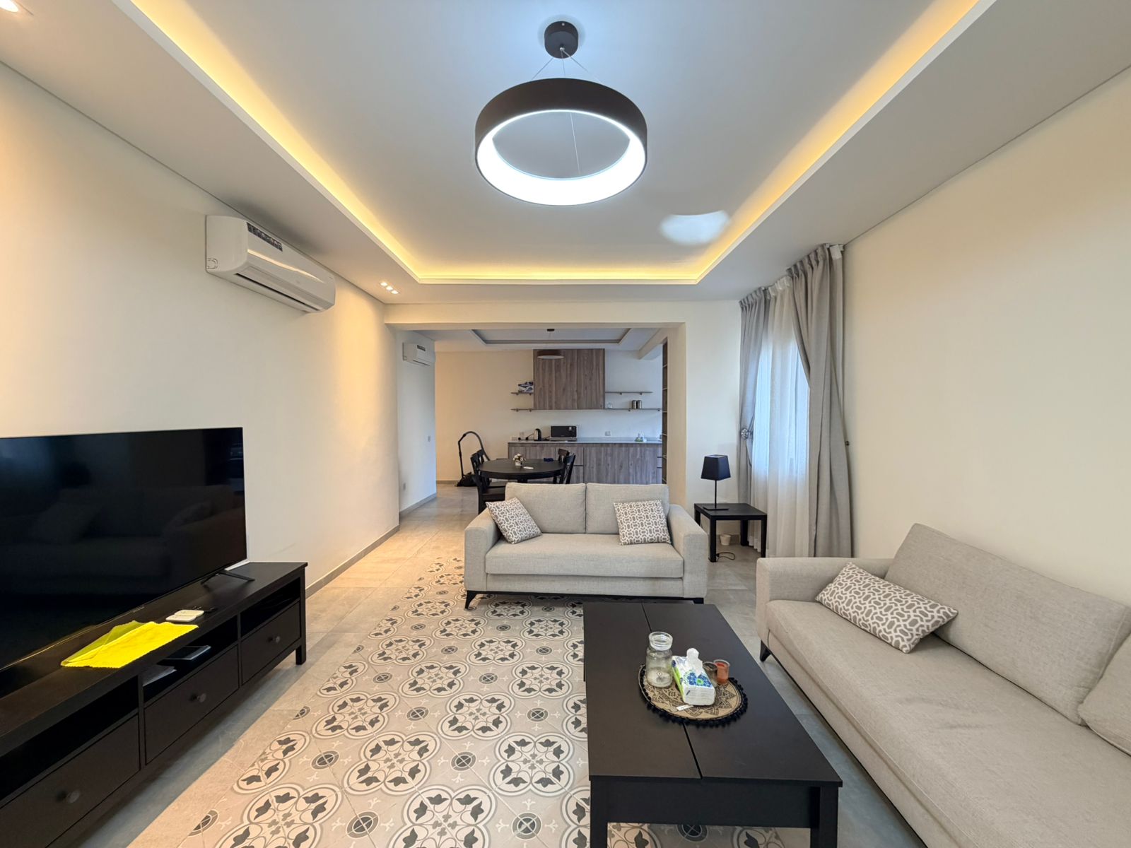 c956dbcb-ba4e-4ff8-a3de-551c7b7b9db1 Bright, modern living room with patterned tile floor, two beige sofas, black coffee table, and a large flat-screen TV.