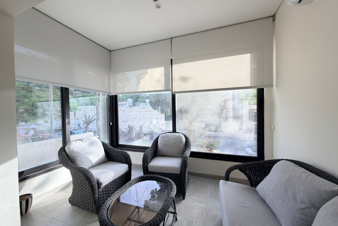 Bright balcony seating area with gray cushioned wicker chairs and a glass table by large windows with roller blinds outside the scene,