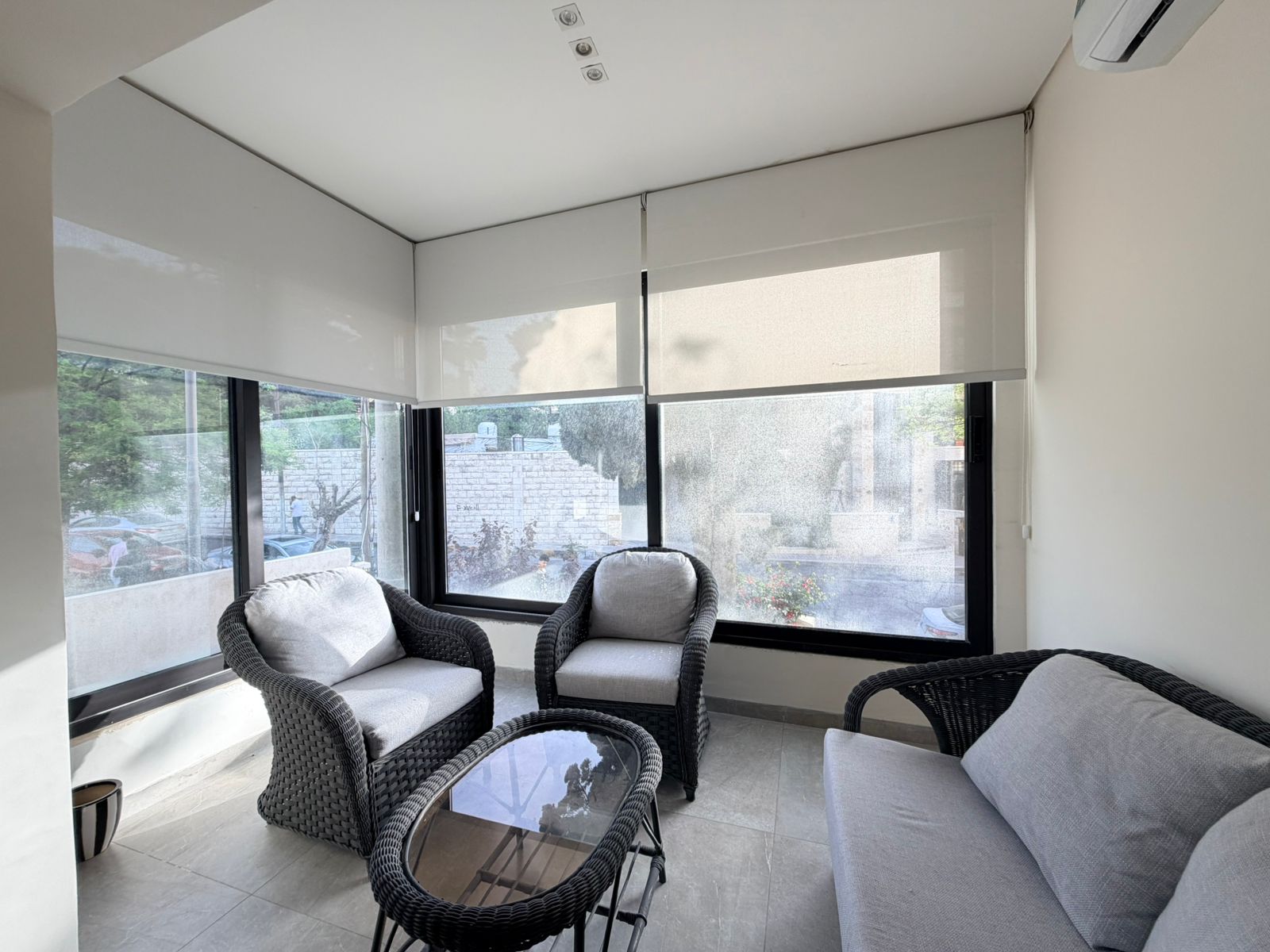 f1b5204a-8282-49dc-bbf2-4a0dd8a87db3 Bright balcony seating area with gray cushioned wicker chairs and a glass table by large windows with roller blinds outside the scene,