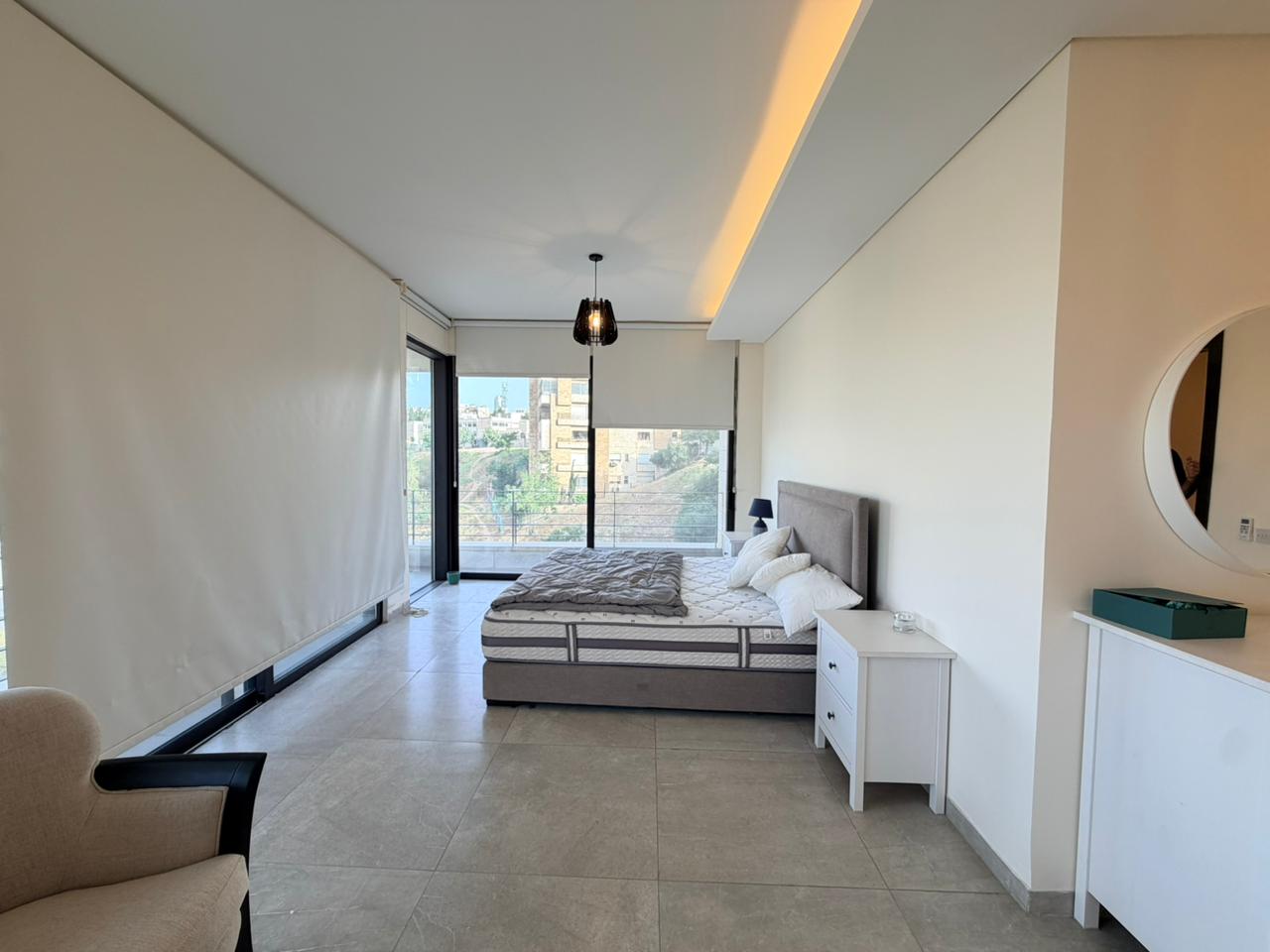 f7319a30-bdf1-45c4-be28-6ddc43760100 Bright minimalist bedroom with a queen bed, gray upholstered headboard, white nightstands, and a city view through large windows.
