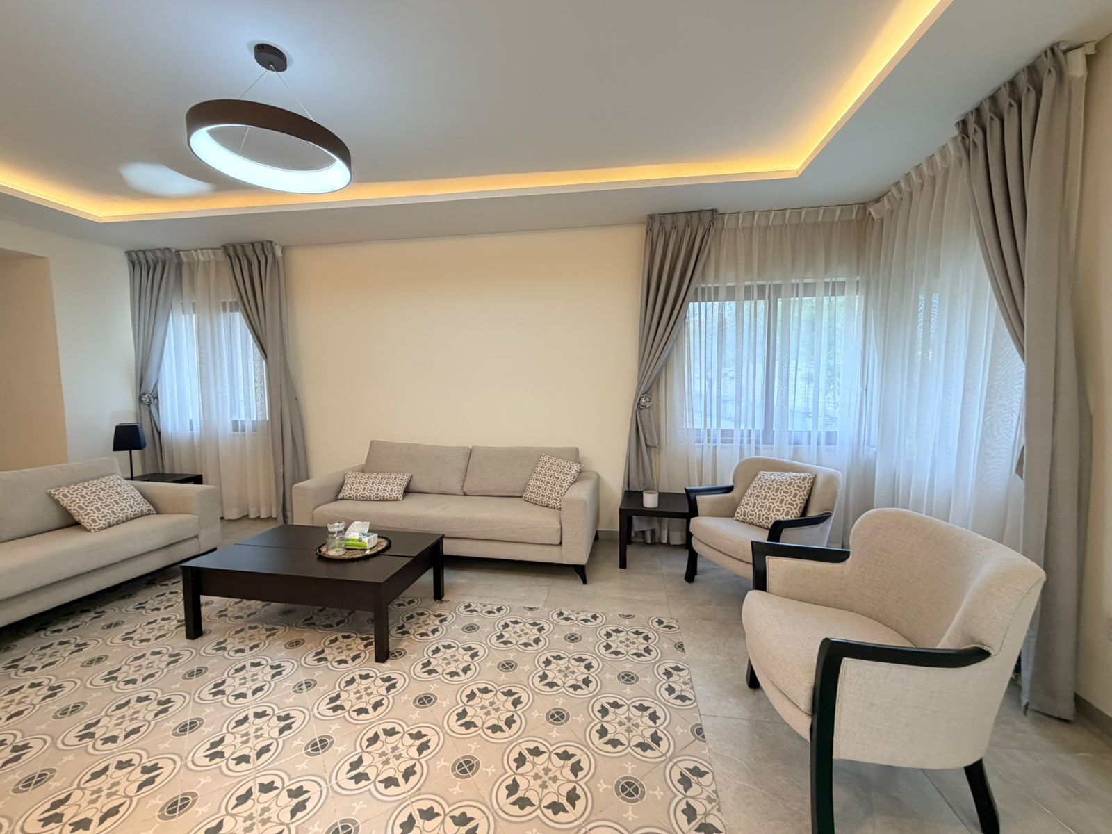 f7467bf0-b040-4e28-a897-51344fccbcc7 Bright living room with beige sofa set, two armchairs, and a dark coffee table on a patterned tiled floor.