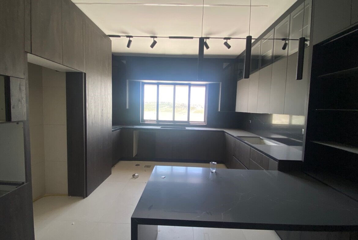 b97945e2-1606-4aba-bbbf-b0d09b139a7b Modern dark kitchen under construction with black cabinets, gray countertops, an island, and a large window at the far end letting in light.