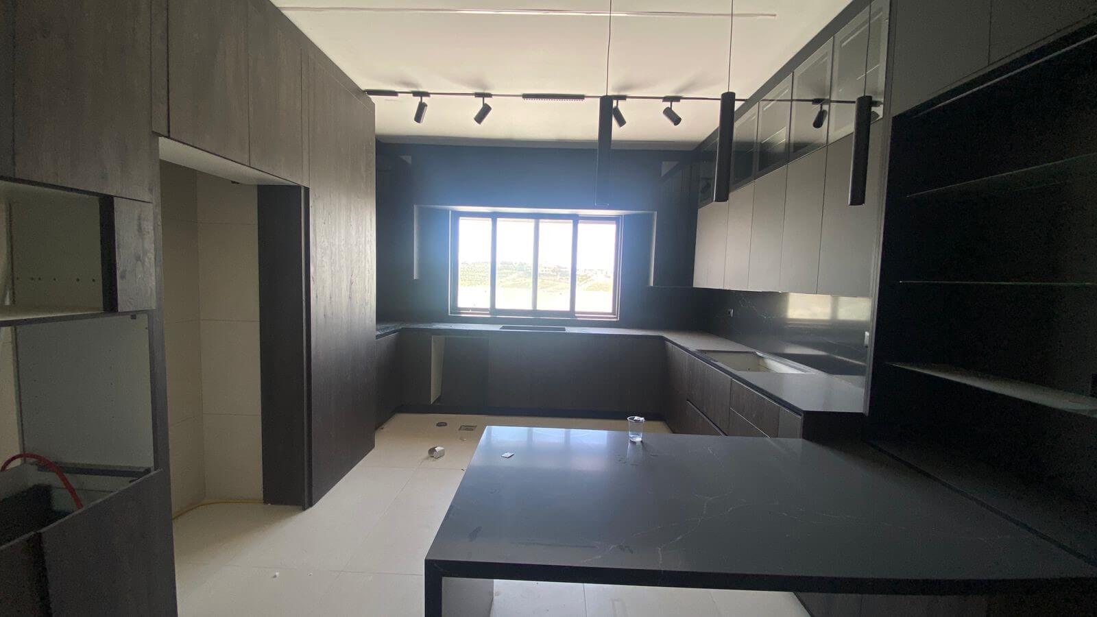 b97945e2-1606-4aba-bbbf-b0d09b139a7b Modern dark kitchen under construction with black cabinets, gray countertops, an island, and a large window at the far end letting in light.