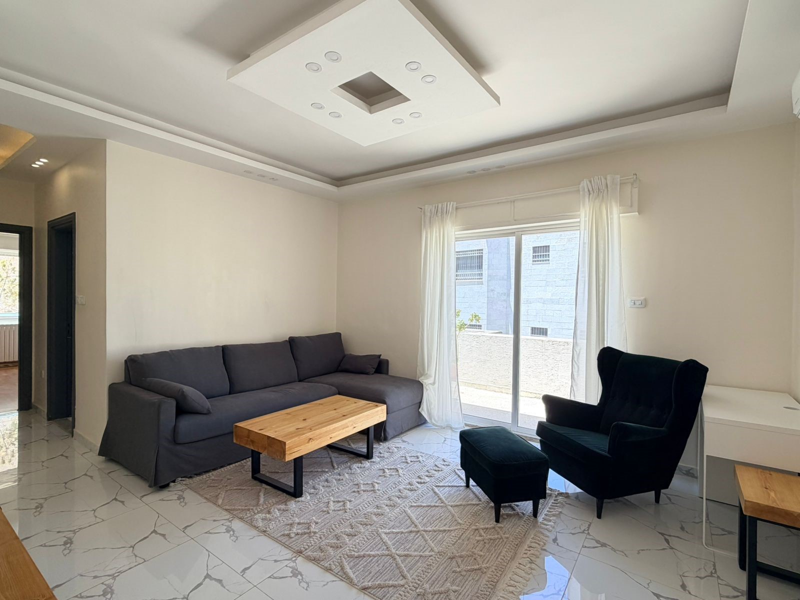 7912d75e-8fc2-43ad-a920-4a307ee2bd1e Living room with a dark gray sectional sofa, wooden coffee table, and a black wing chair facing a sliding glass door with white curtains.