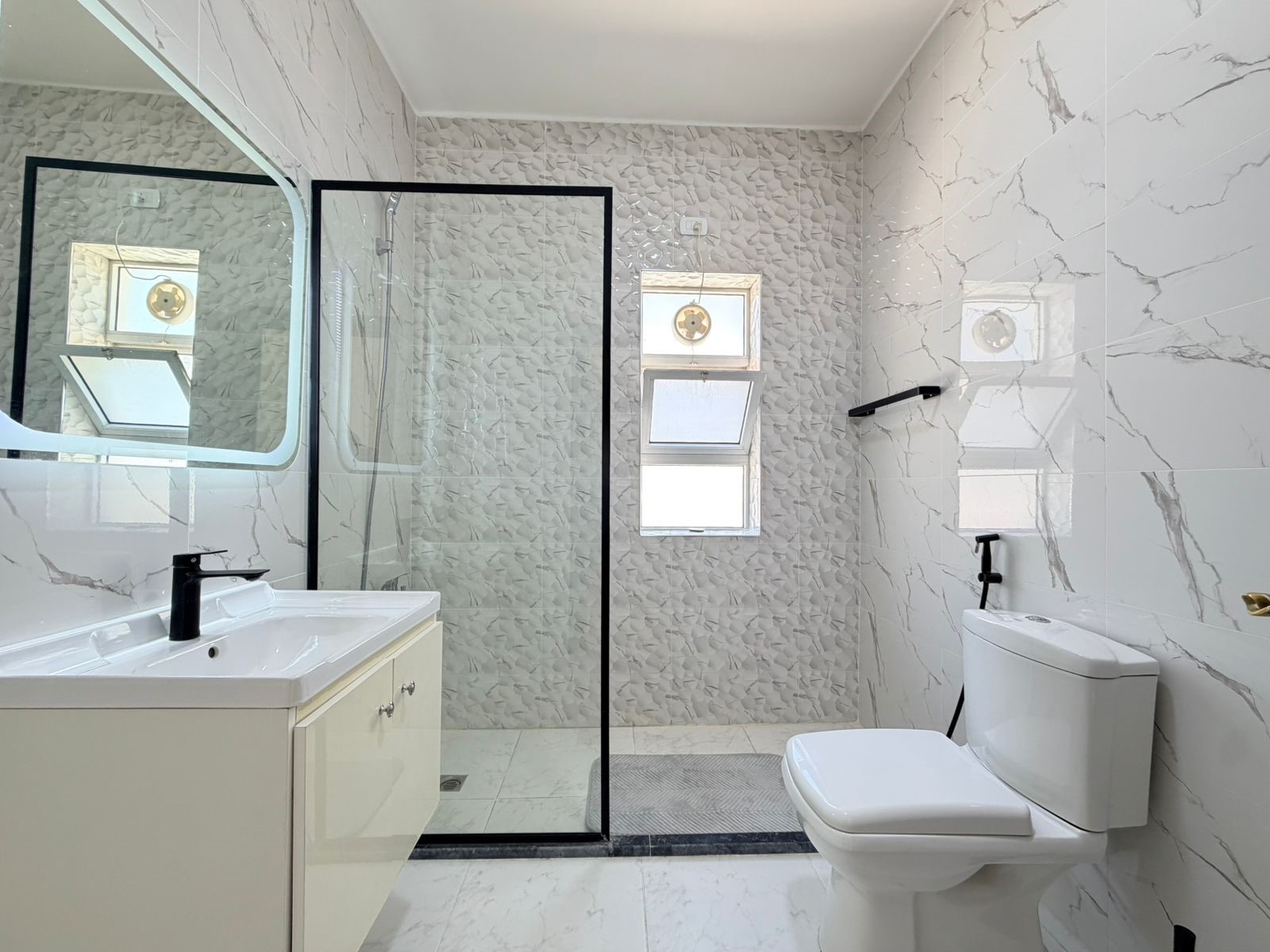 7f518a13-c5ce-4a4e-b33c-55ea359f5d56 Modern bathroom interior with a glass shower enclosure, white vanity, and a toilet in a marble-tiled room