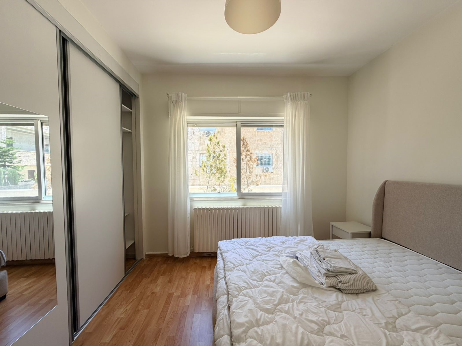 85e65c86-2fdd-492d-898b-c262a03dcbcd Bright bedroom with a large sliding-door wardrobe on the left, a white quilted bed on the right, and a window with sheer white curtains opposite the bed, radiator beneath the window.