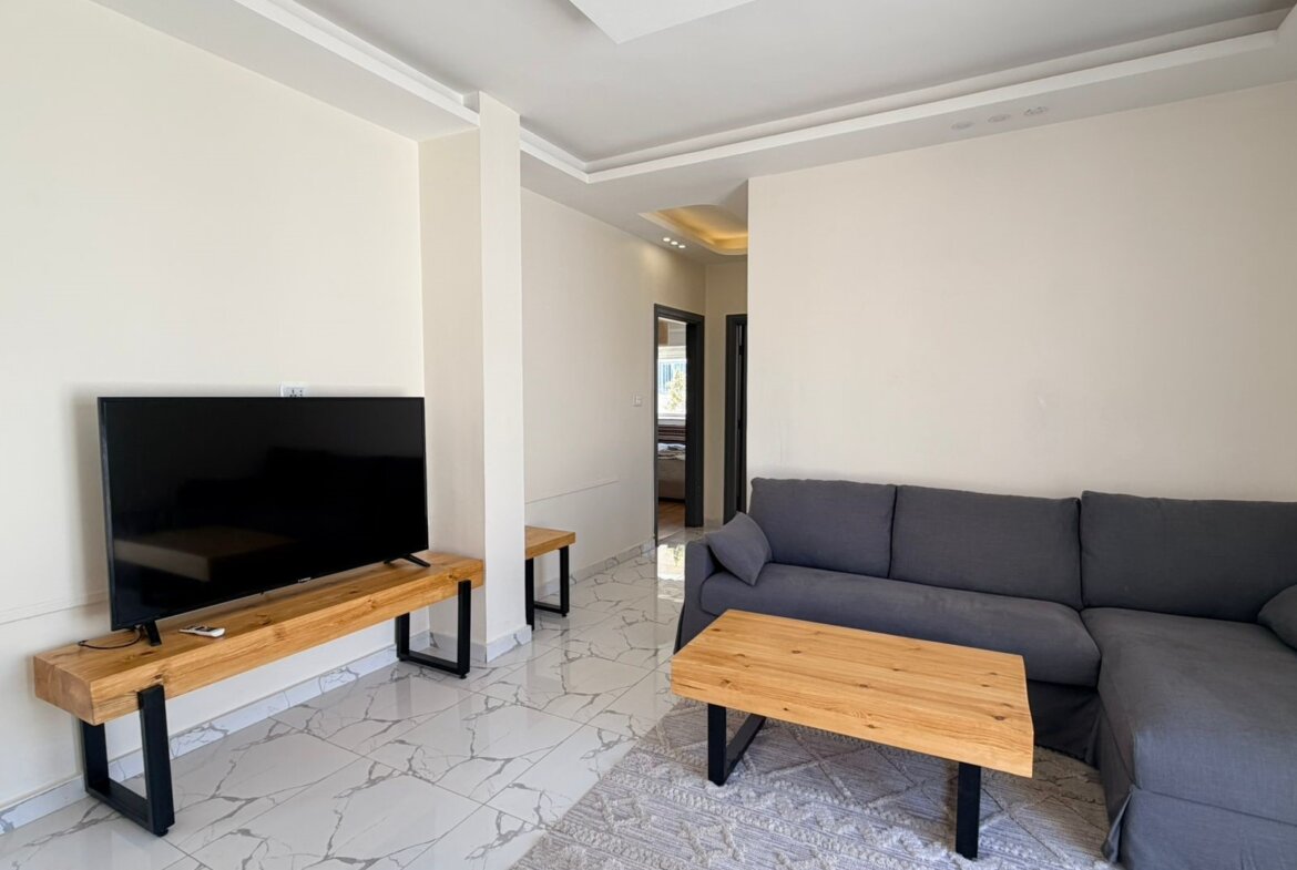 Television on a wooden console with black legs, facing a gray sectional sofa in a bright living room. (Informative)