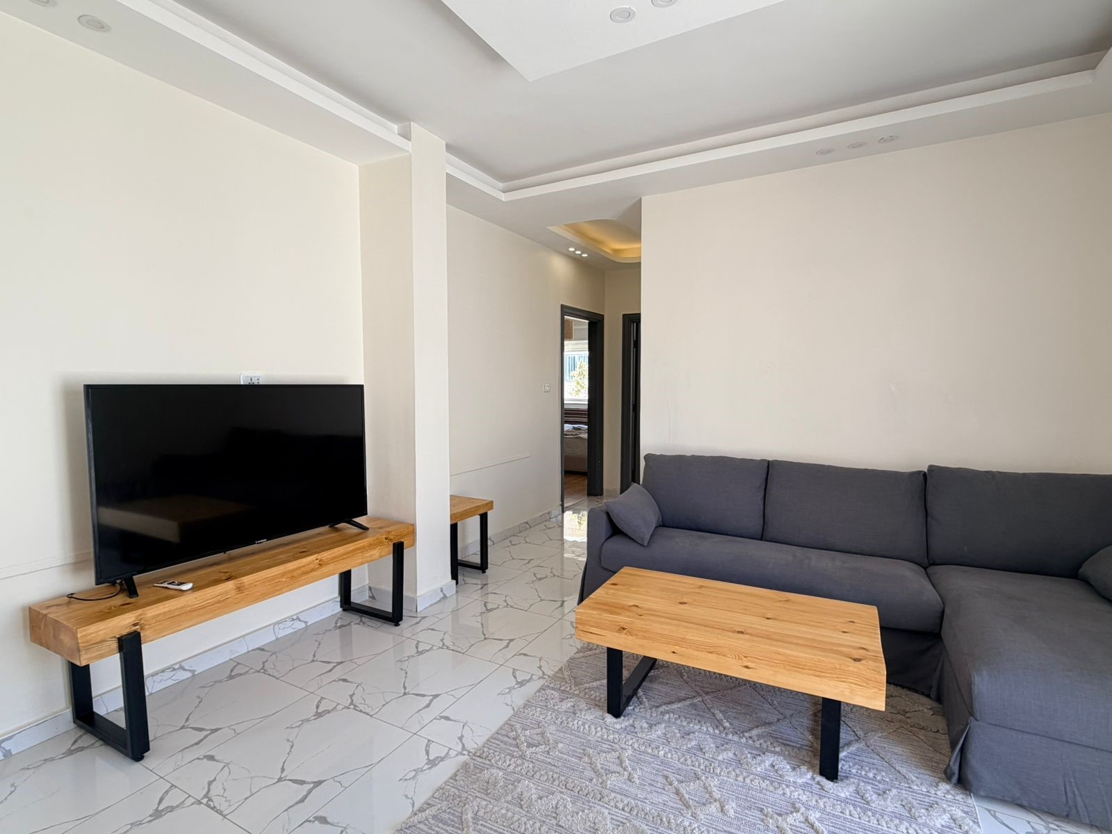 9475b344-3373-43ce-8cf4-9bc53a52756a Television on a wooden console with black legs, facing a gray sectional sofa in a bright living room. (Informative)