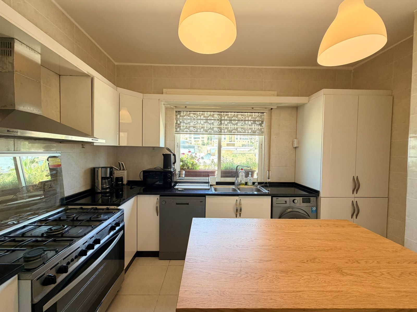 9a271c92-a0fd-44e5-81a5-a0e4d964e67f Modern kitchen with white cabinets, stainless steel appliances, black countertops, and a wooden island in the foreground; window overlooks a garden.