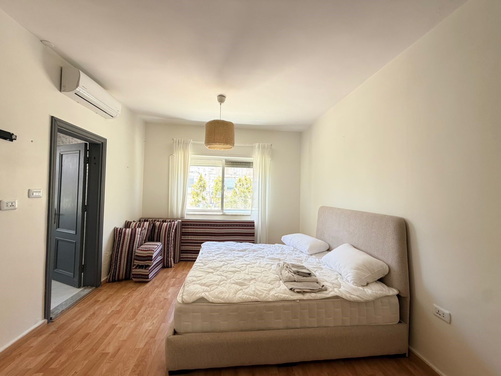9fe7ddda-c909-4c92-94c5-a12612f5c543 Bright bedroom with a large bed and white bedding, beige headboard, and folded towels at the foot of the bed; window with sheer curtains at back.