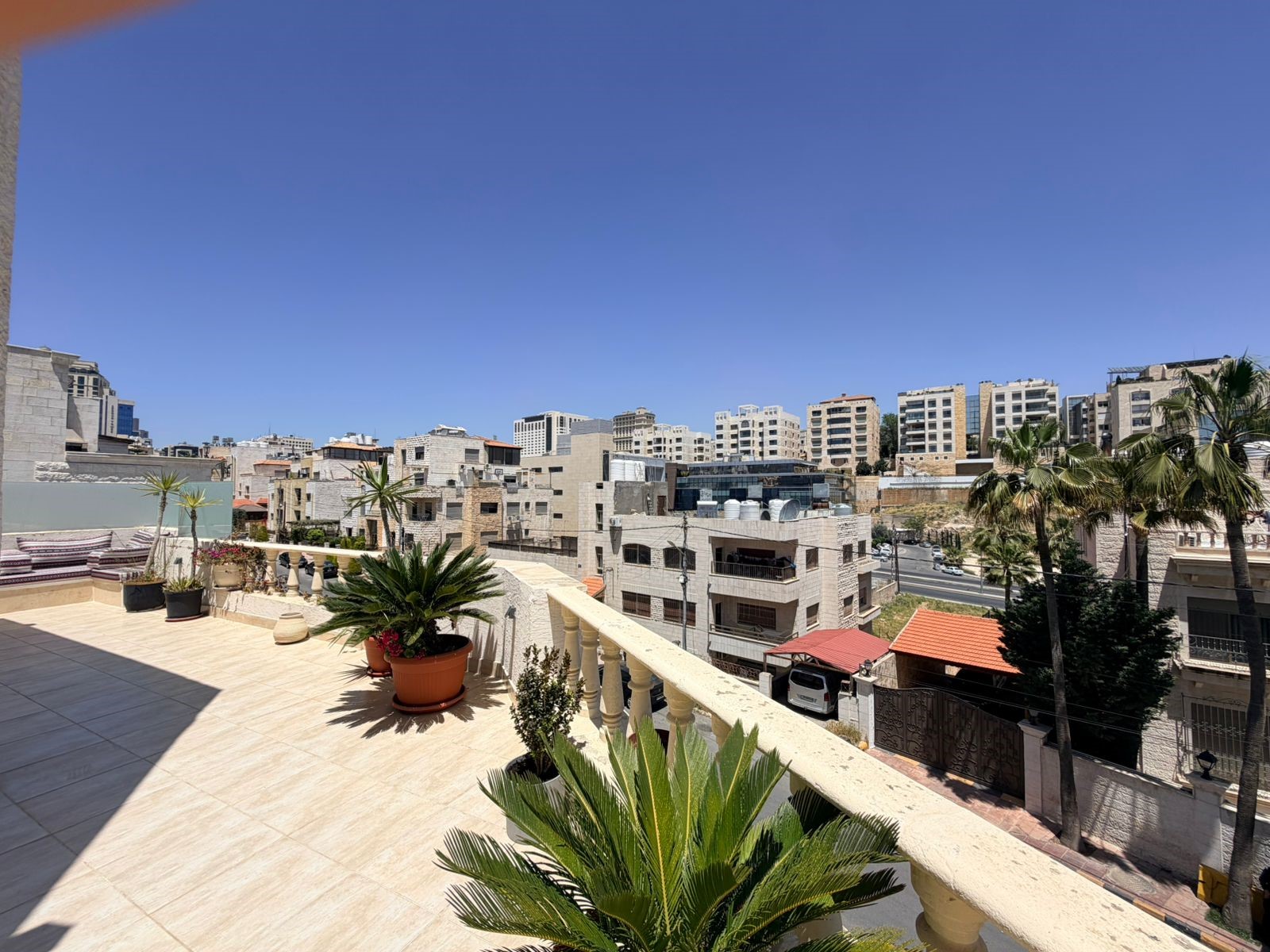 c27007e1-070e-42cb-a9cc-ad04d44b3f61 Sunlit rooftop terrace with potted plants overlooking beige city buildings under a clear blue sky, palm trees in view.
