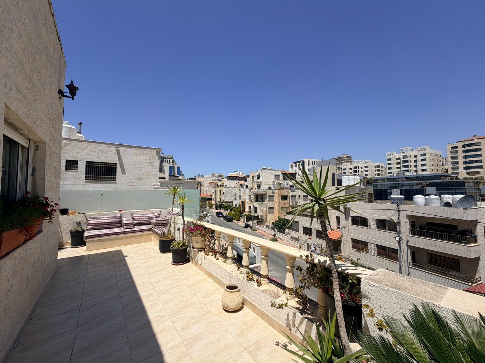 d5251afe-345f-4263-9be3-060081d6f84b Sunlit rooftop terrace with a cushioned seating bench, potted plants, and a balustrade overlooking a city of beige stone buildings under a clear blue sky.