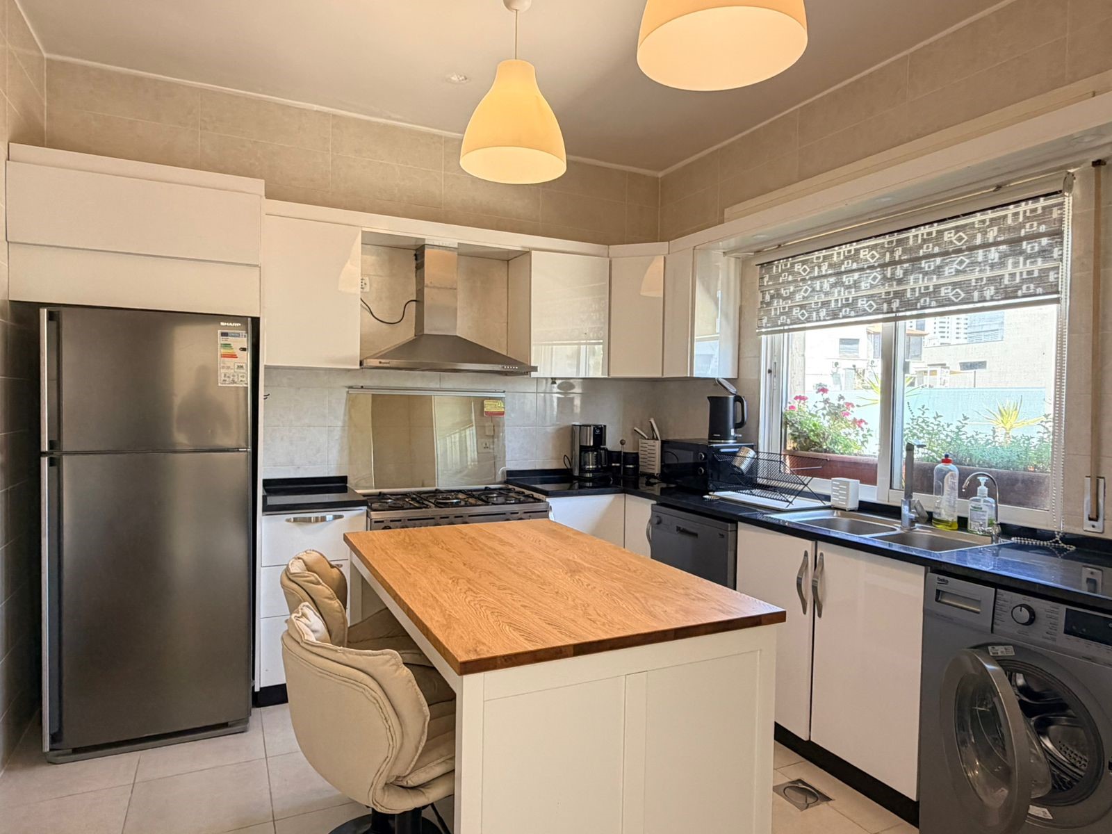 e8c9bd1b-efbc-43f6-8a52-f3ba3c4bda70 Modern kitchen with stainless fridge, white cabinets, and a wooden island; pendant lights above and a large window with a patterned blind to the right