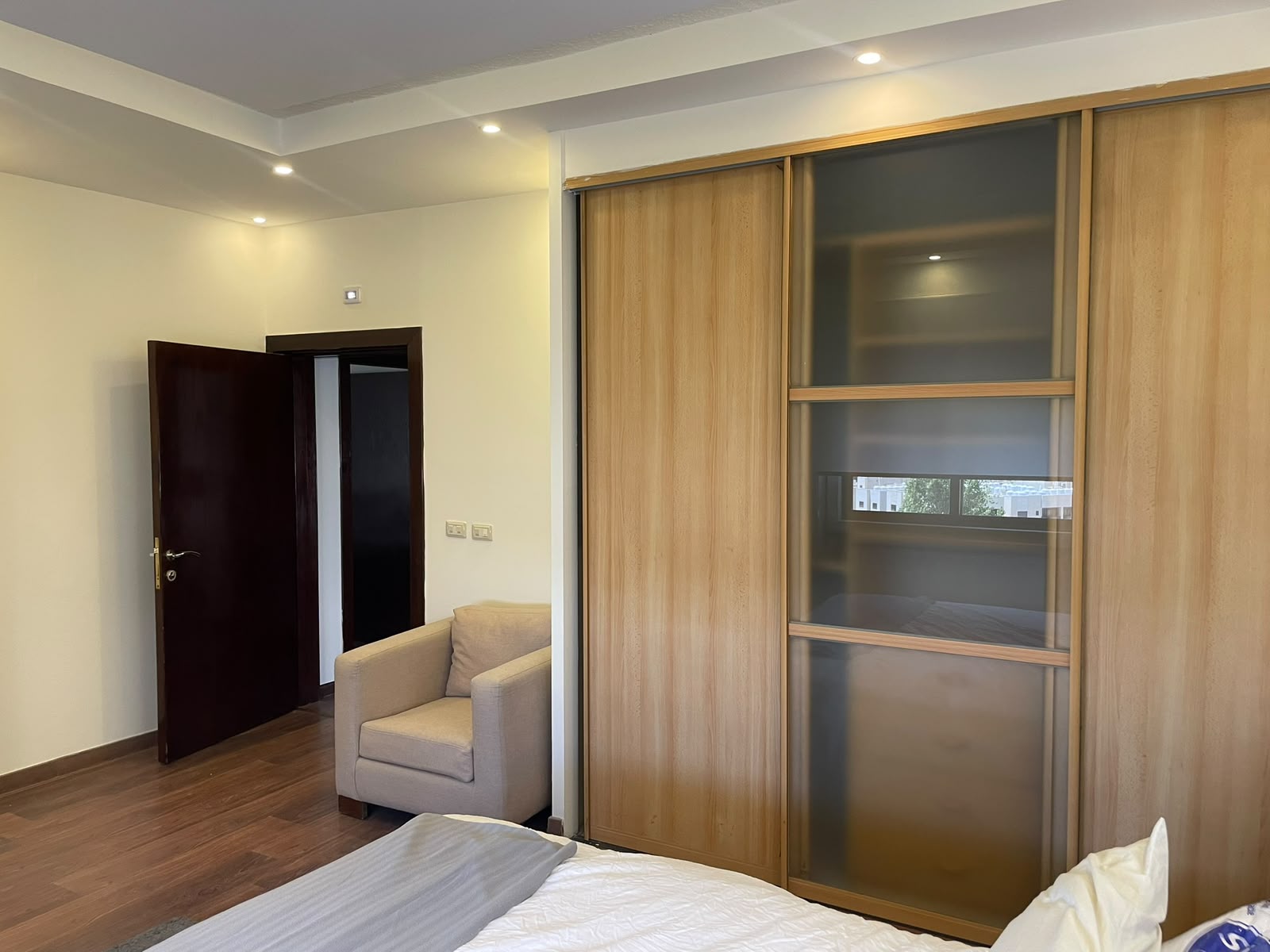 10c4609f-bddb-4f4a-823d-2dc988a993d9 Bedroom with a wooden sliding wardrobe featuring frosted glass panels, a beige armchair, and an open dark door to a hallway in the background.