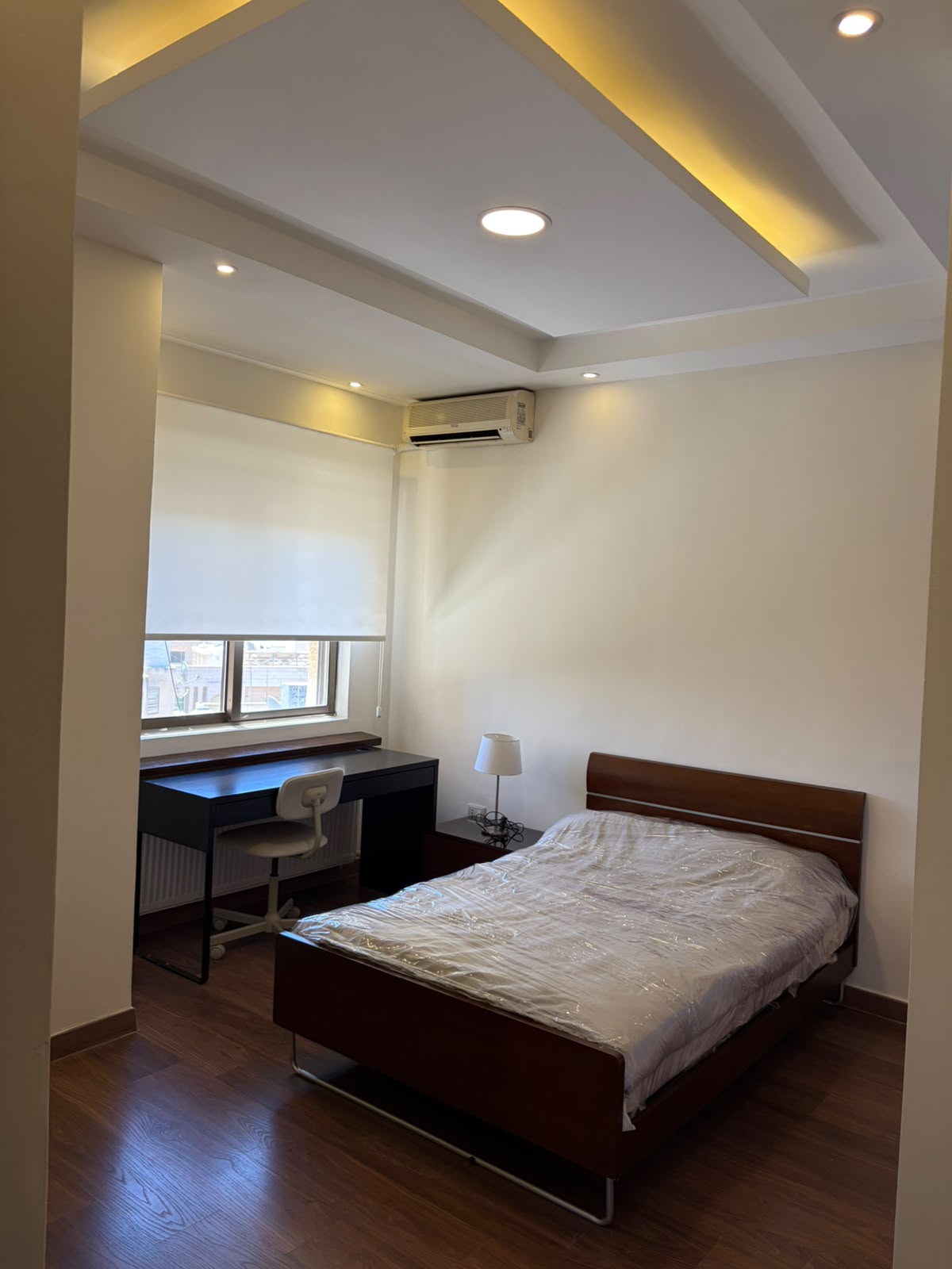 13f6d7a6-9025-4312-b5a6-f8d7f4292f8c Minimalist bedroom with a single wooden bed, nightstand lamp, and a dark desk by a window with blinds; warm ceiling lights.