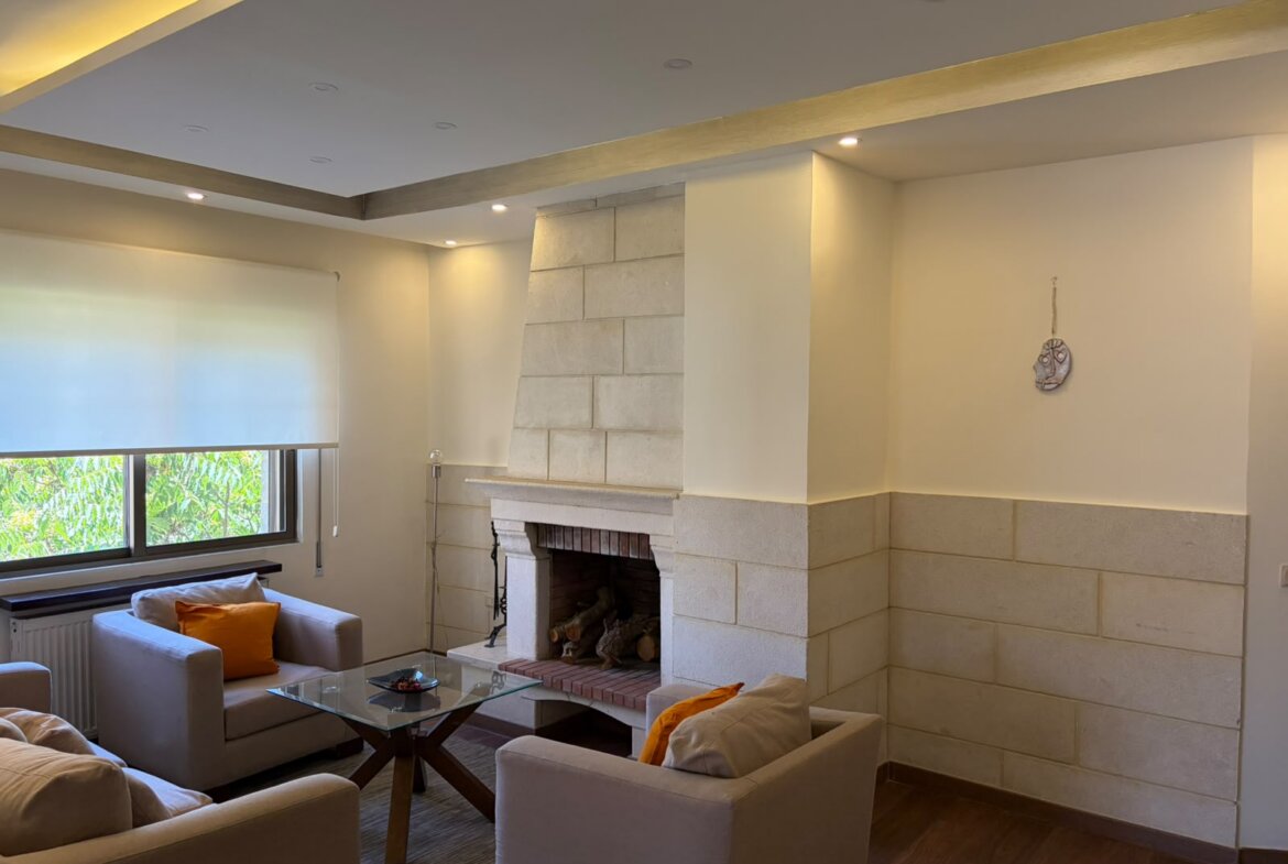 Cozy living room with beige sofas around a glass coffee table and a stone fireplace beneath a recessed ceiling with warm lighting and orange cushions on the chairs while a window casts natural light.