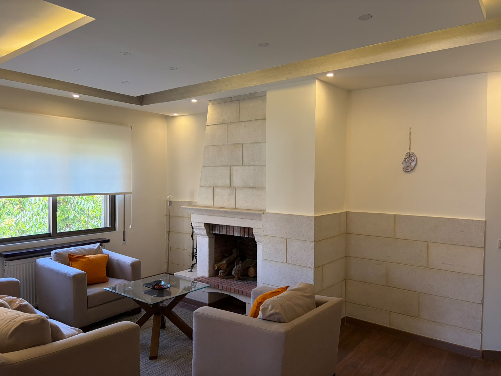268164bf-daaf-4b08-820b-862586803fe2 Cozy living room with beige sofas around a glass coffee table and a stone fireplace beneath a recessed ceiling with warm lighting and orange cushions on the chairs while a window casts natural light.