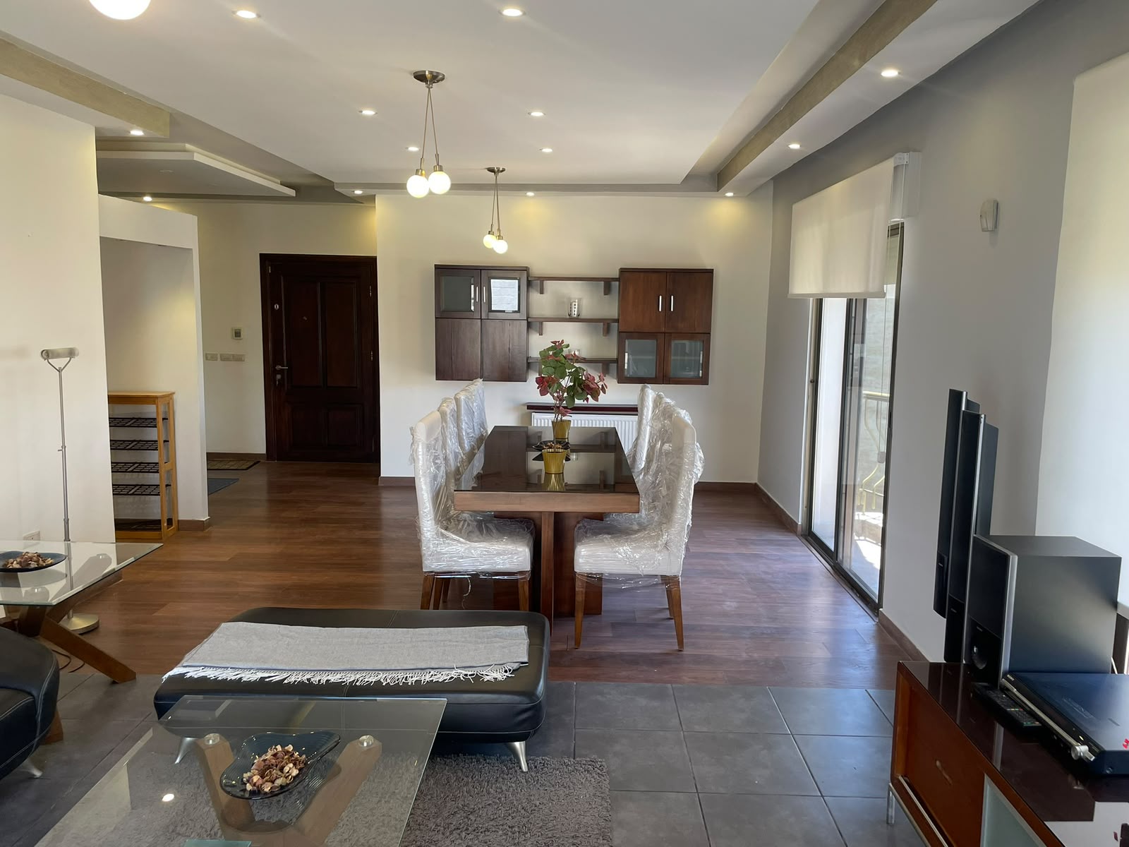 4bb763b6-3936-485f-addf-3972fef790d1 Bright, modern dining area with a glass table and six plastic-wrapped chairs, a centered plant, and a dark wood wall unit. Floor-to-ceiling windows to the right and a TV on a stand nearby.