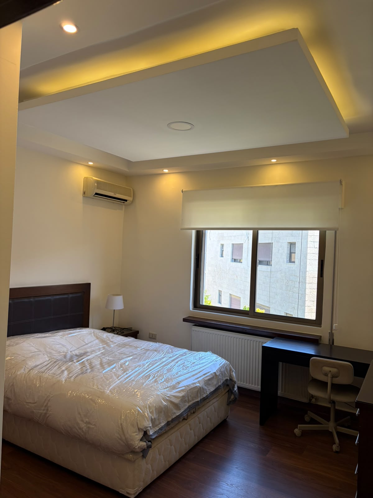 61132307-9e4d-440d-add6-d698f48a1491 Bedroom with a large bed and padded headboard, nightstand lamp, and a desk by the window.