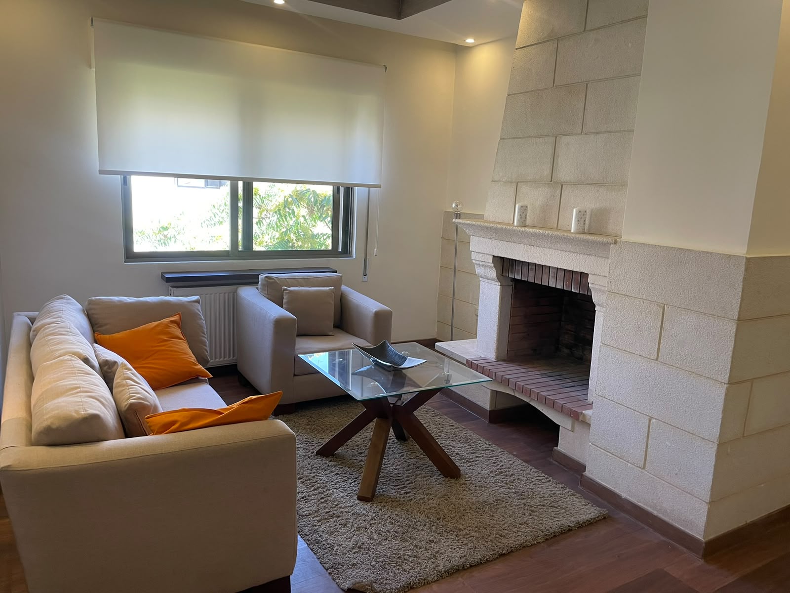 834691b7-9f08-4928-829e-bfce9f8a5da1 Cozy living room with a beige sectional, orange cushions, a glass coffee table, and a stone fireplace. The window has a white shade and warm lighting.