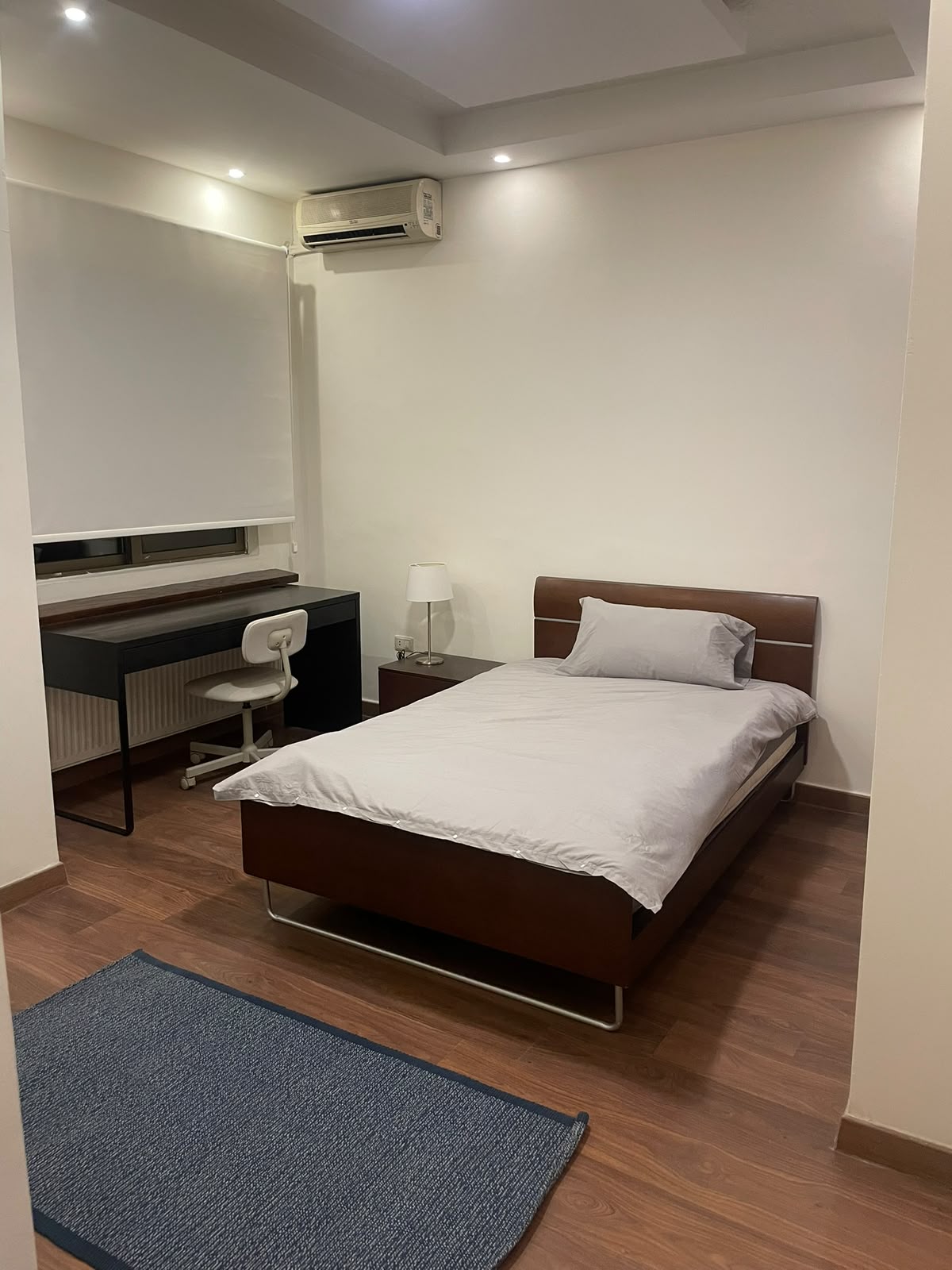a32b9e75-4ebf-4702-9ea0-9aa172a5abc1 Single bed with grey bedding and brown headboard in a minimalist room, a desk and chair on the left and a window with blinds above.