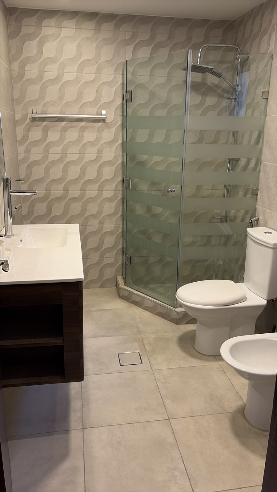 afda4eb3-2044-4026-a6f1-8da2457aacd3 Modern bathroom with wavy-patterned gray wall tiles, a glass shower enclosure with frosted horizontal bands, a white toilet and bidet, and a dark wood vanity with a white sink.