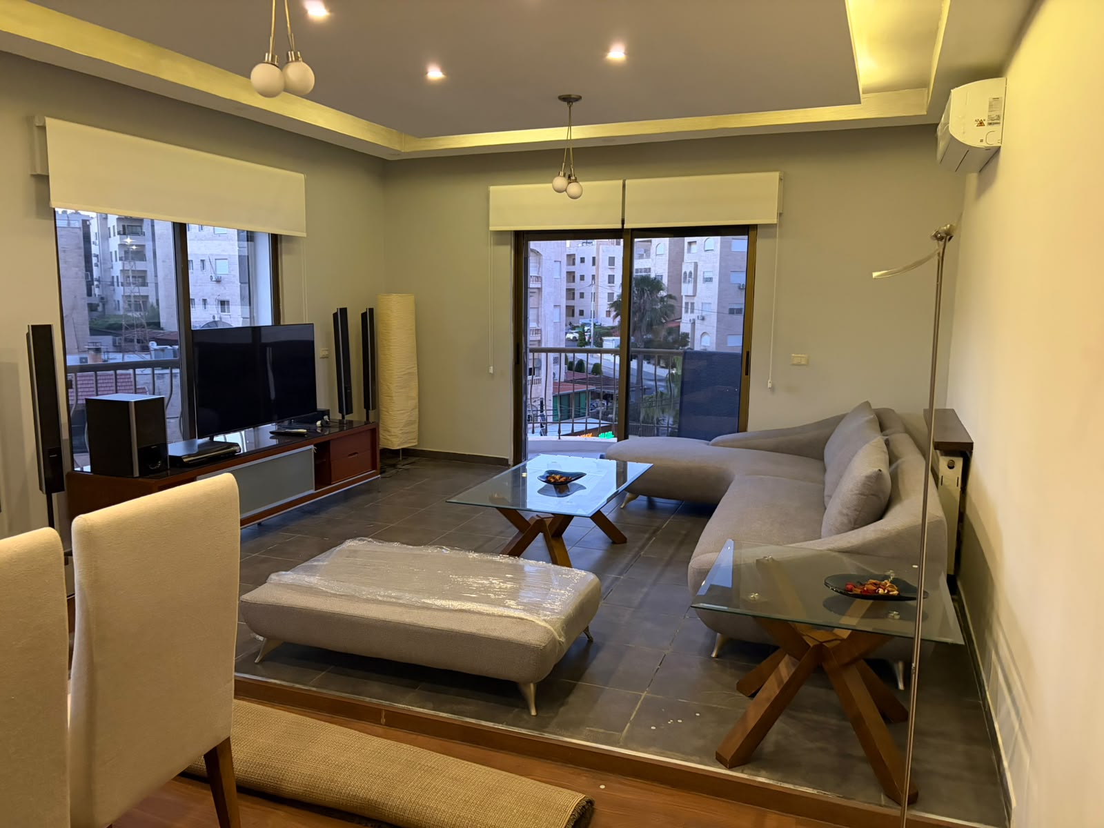 b36e10e3-915f-42f3-9bd5-c5f36e055950 Modern living room with gray sectional sofa, glass coffee table, and a TV setup facing a balcony view of buildings outside the windows.