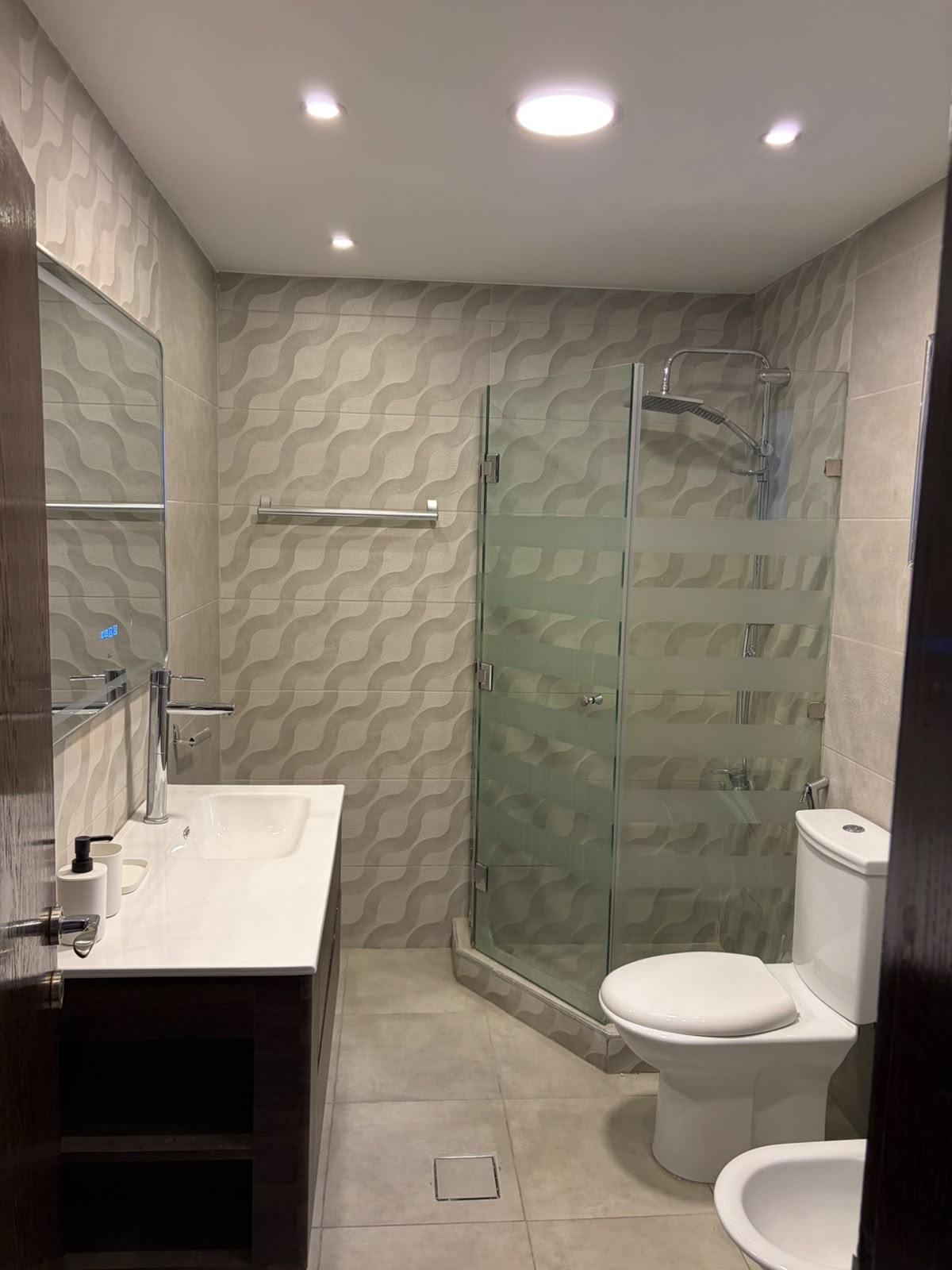 b946420f-061a-4792-984c-4b03d3bce6fe Modern bathroom with glass shower enclosure, white vanity, and patterned gray wall tiles.