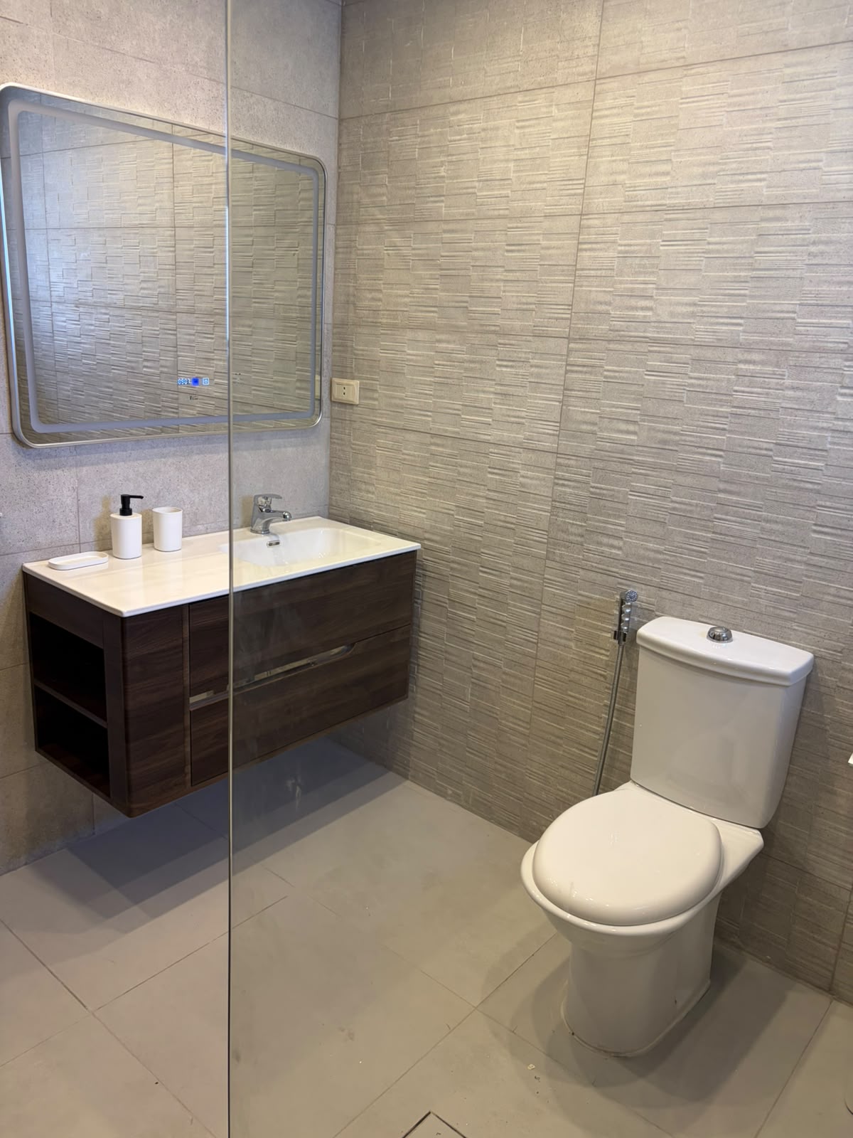 bf82ff4a-ba66-4b8d-a2ed-339695cb052a Modern bathroom with a glass shower divider, textured wall tiles, a dark wood vanity with white countertop, and a white toilet.