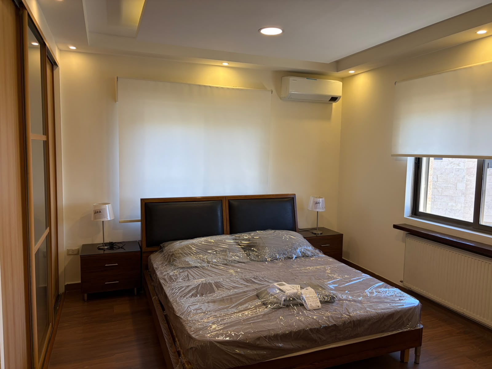 c9331651-cc83-4bf0-8f51-51be02c2edd3 Bedroom with a plastic-covered bed, two wooden nightstands, and soft warm lighting from recessed ceiling lights.