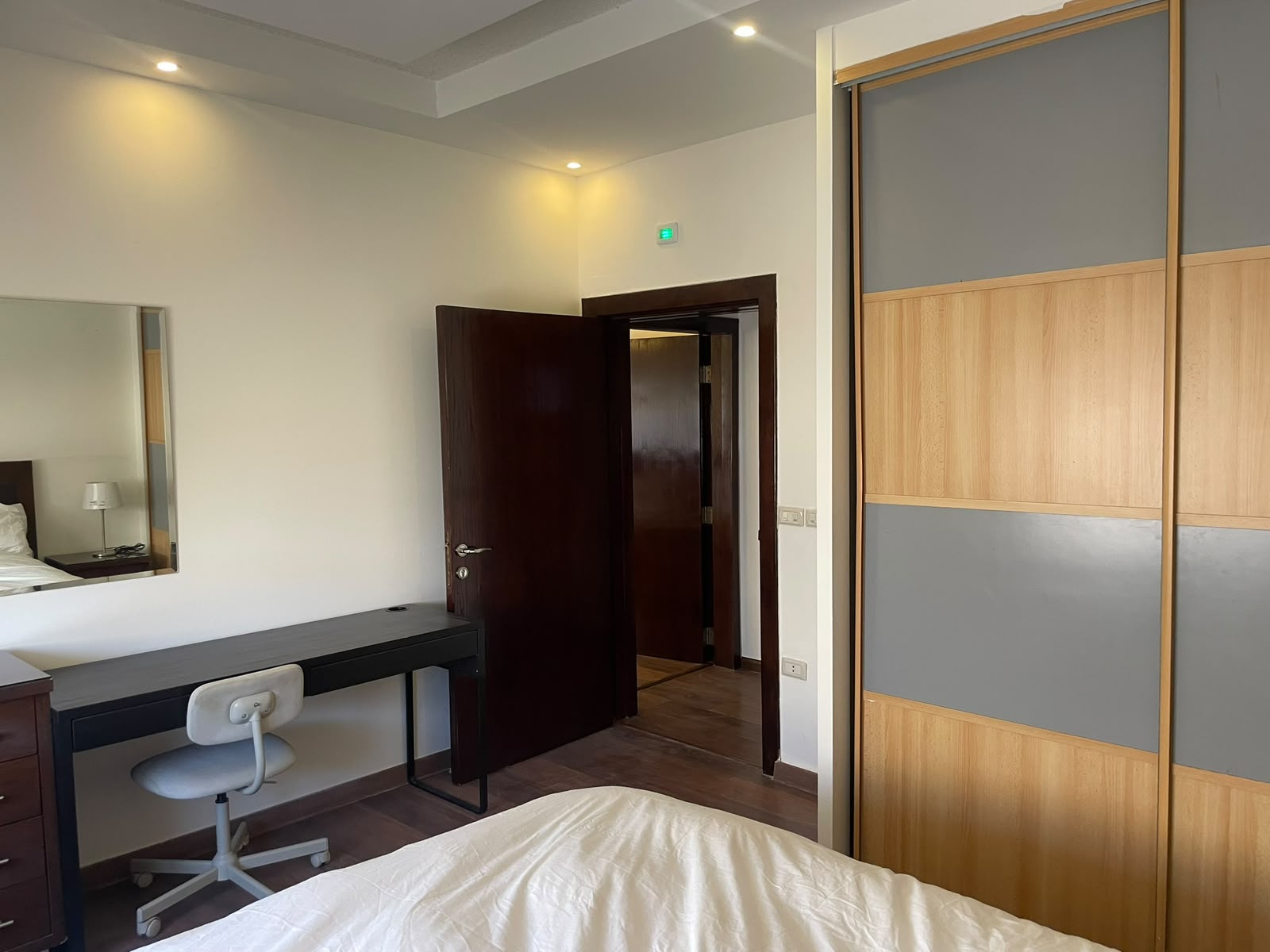 fcf92212-e03d-41e4-aa0a-20d6d7c69bd6 Bedroom with white bedding in foreground, dark desk and swivel chair to the left, open dark wood door, and a tall two-tone wardrobe with wood and gray panels on the right.