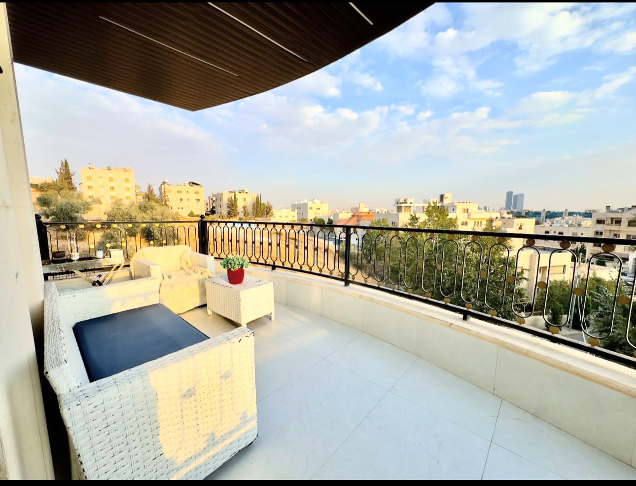 190fb2eb-fb2e-437f-a1e5-0cb2d0819d58 Urban balcony with white wicker seating, navy cushions, and a small potted plant, overlooking a cityscape.