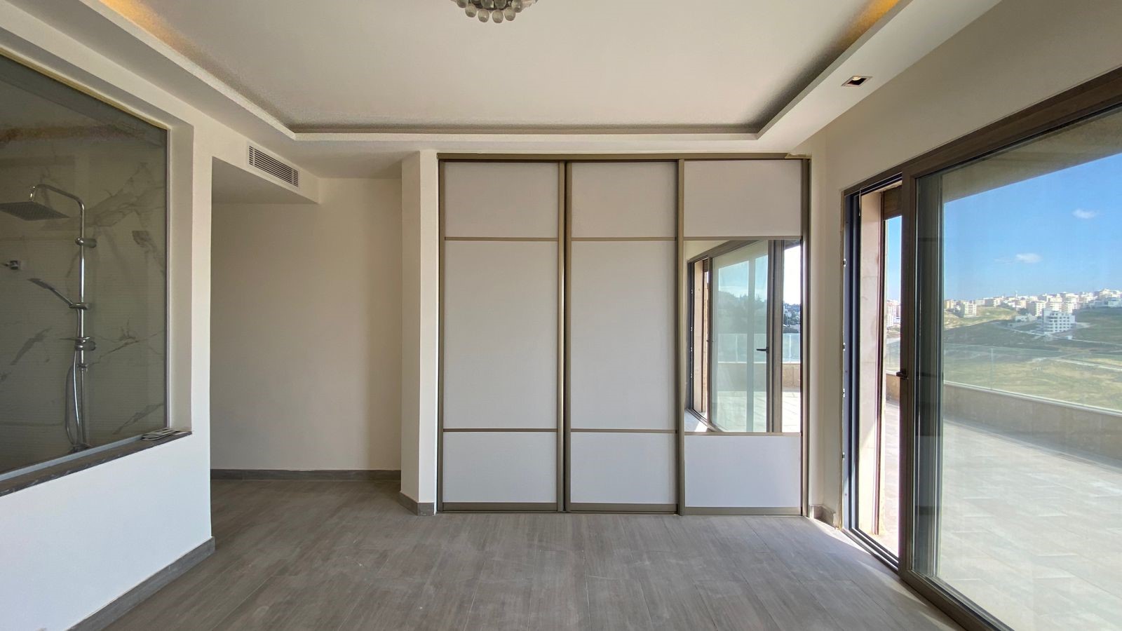 02183326-7967-42dc-b0a9-1f7bbefe5678 Empty modern living area with a large sliding-door wardrobe, a glass-walled shower on the left, and sliding glass doors to a balcony with a city view.