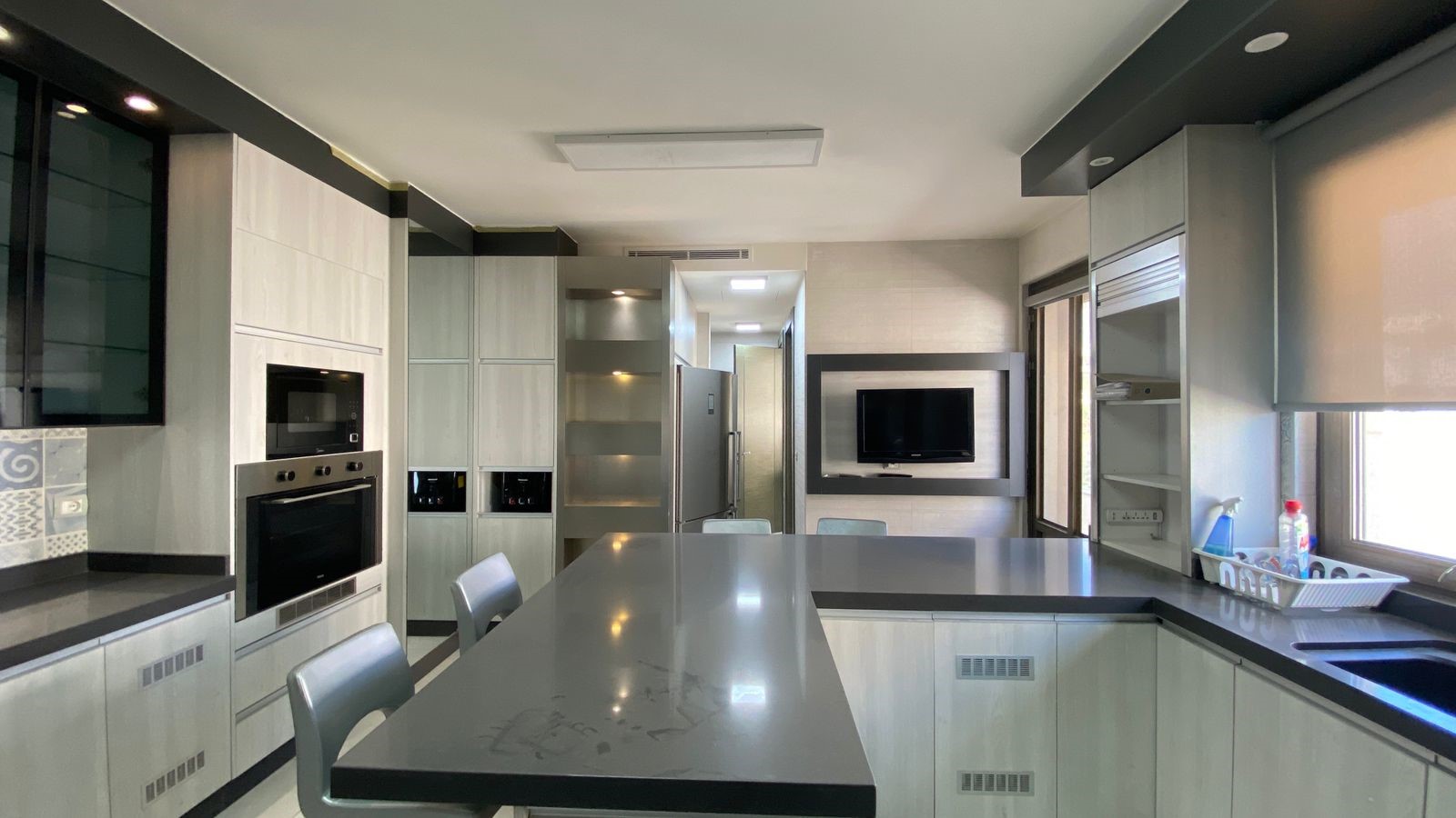 0f275633-e1d1-43a6-8ea8-dd1b32758593 Modern kitchen with an island, stainless steel appliances, and a wall-mounted TV on the back wall.