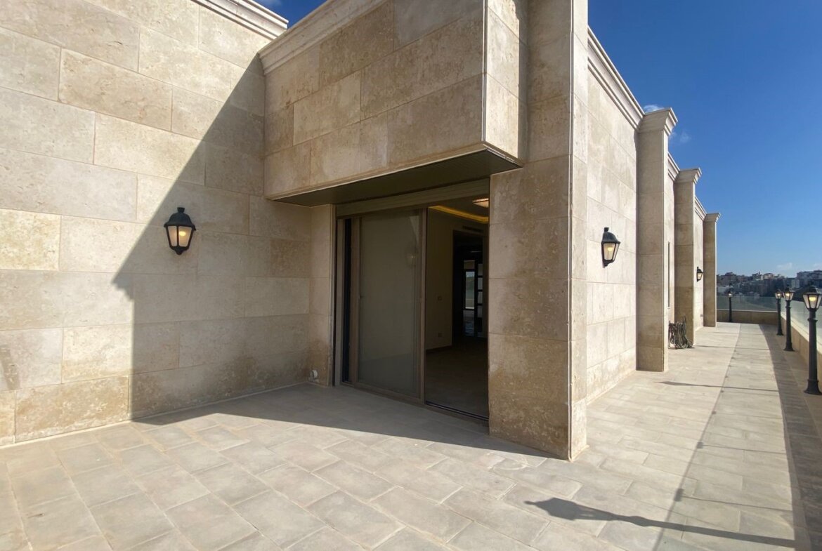 Beige stone rooftop terrace with a sliding glass door, wall lanterns, and a glass railing overlooking a city skyline under a blue sky.
