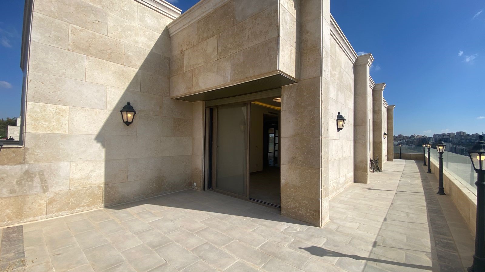 0fb05ea1-38d9-42d3-9c98-54693b0e5ea8 Beige stone rooftop terrace with a sliding glass door, wall lanterns, and a glass railing overlooking a city skyline under a blue sky.