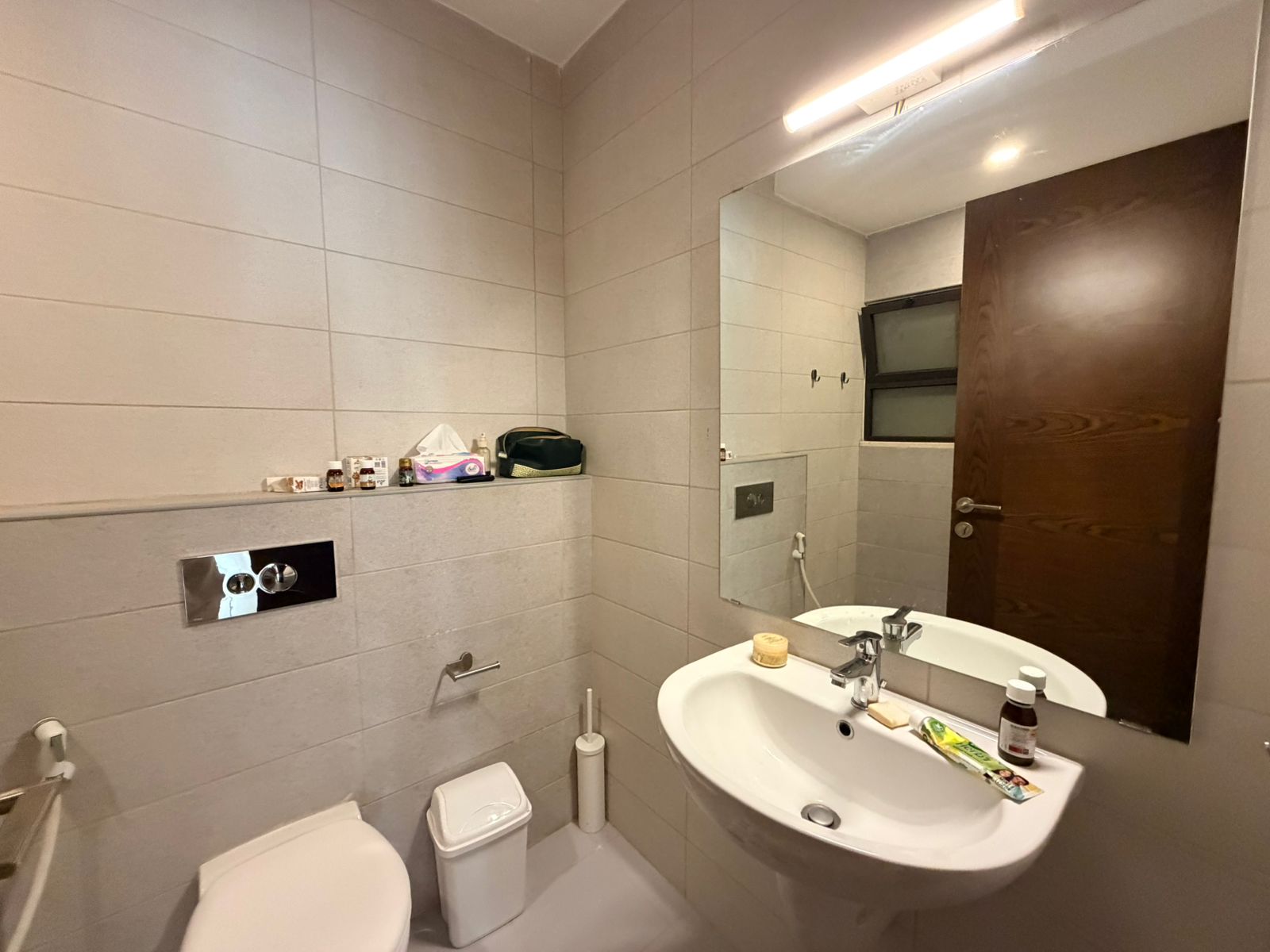 131caff3-4f90-4f3c-b85f-42fc5506ccbb Small modern bathroom with wall-mounted sink and large mirror, toiletries on a ledge, toilet, and wooden door nearby.