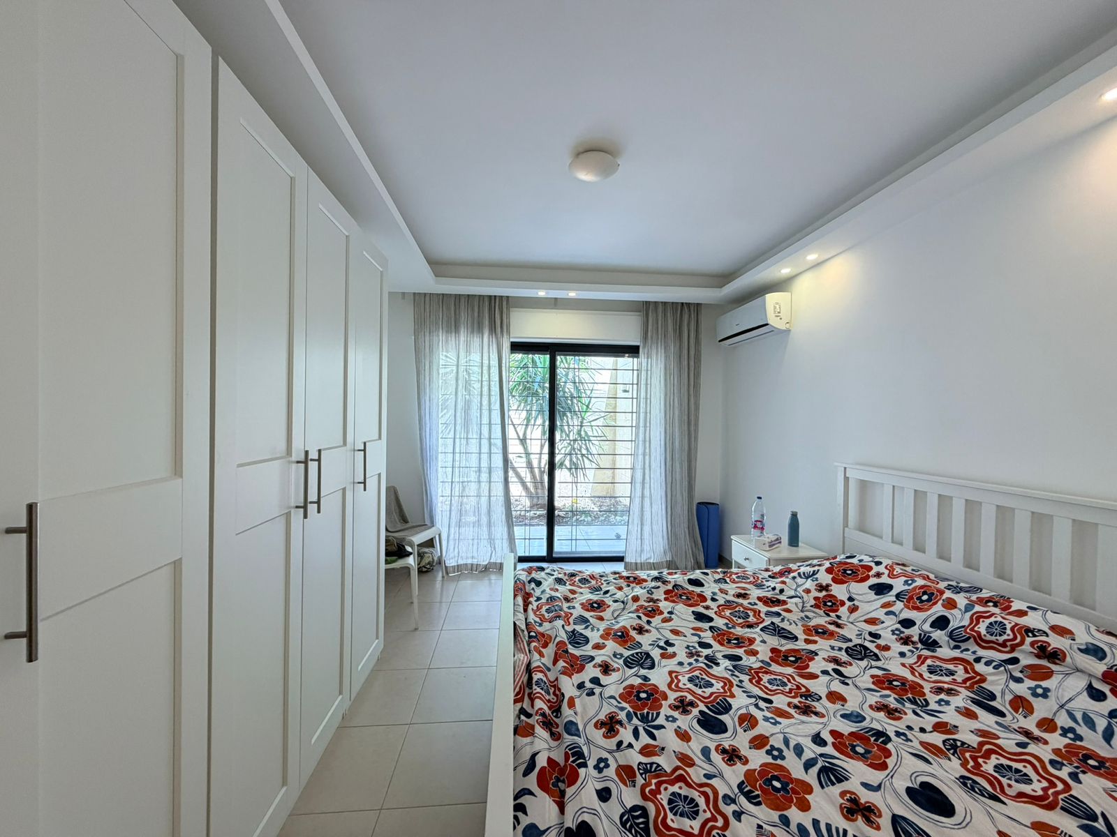 1eb431b7-2053-42e1-8421-515bce6a51ac Bright white bedroom with tall wardrobes on the left, a white bed with a red-blue floral bedspread, and a glass door at the far end.