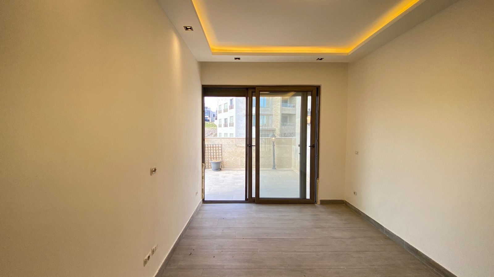 1ebf226c-1003-411d-8bff-844f317d3f6c Empty rectangular room with light beige walls, wood-look flooring, and warm recessed ceiling lights; sliding glass doors open to an outdoor patio.