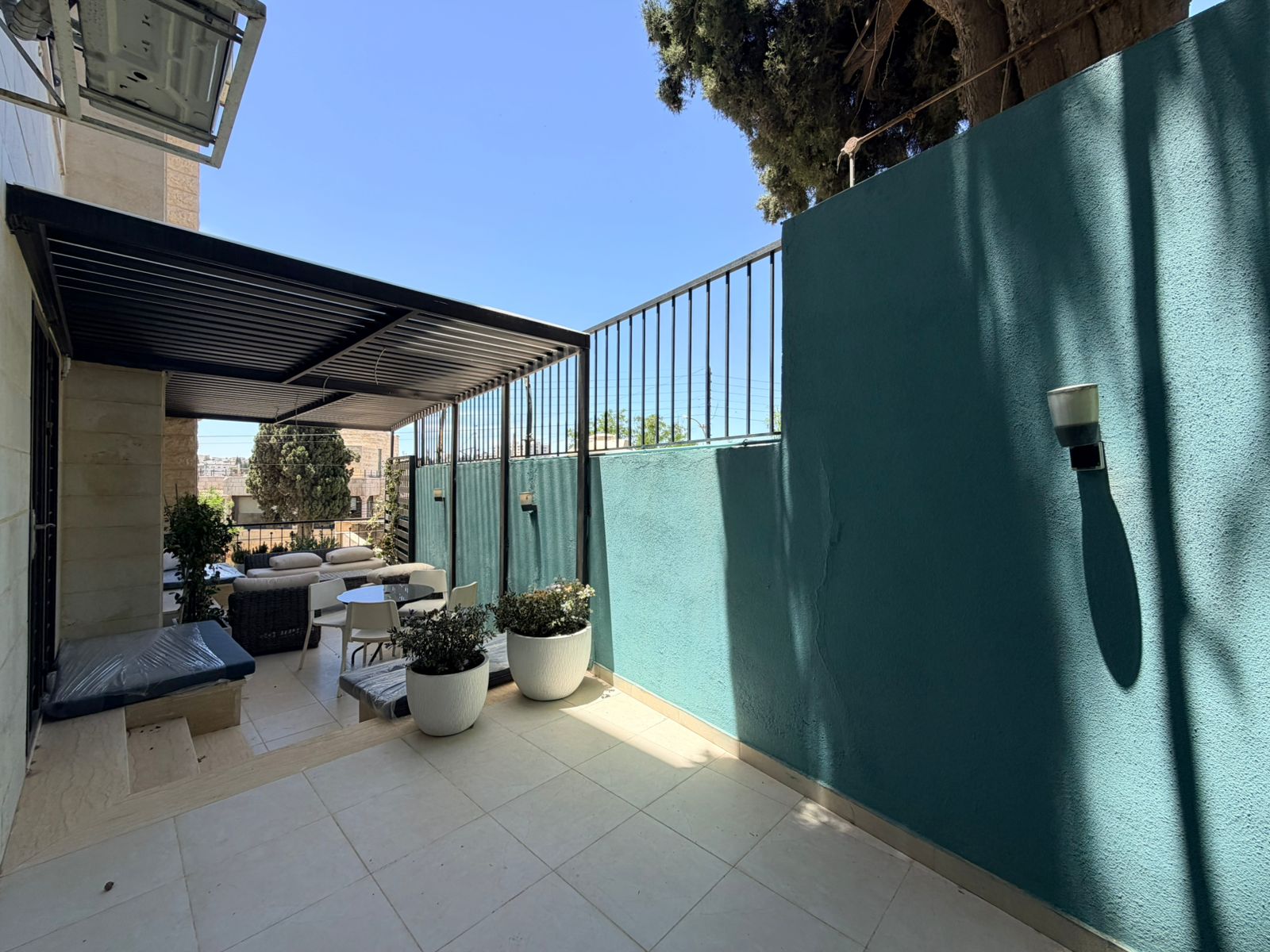 281ac94f-cffa-41ad-b01a-ec61634b70a0 Outdoor patio with white seating, potted plants, and a teal surrounding wall, under a metal-framed shade structure in bright daylight, city beyond.
