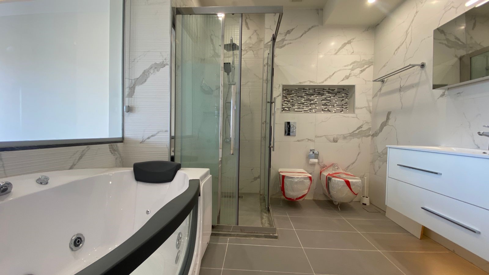 37e4c383-3b7f-42d0-87d2-0fc310fa2d9e Modern bathroom with white marble walls, glass shower enclosure, and a large tub with a headrest; white vanity on the right.