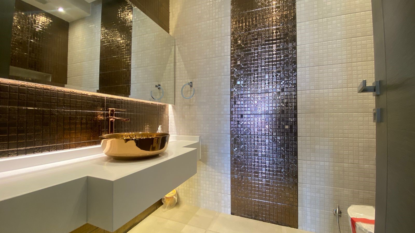 479dc1a4-0d60-4d3f-83d8-4416f64984f5 Modern bathroom with a gold vessel sink on a white floating vanity, brown mosaic tile backsplash, and a vertical brown tile stripe in cream walls.