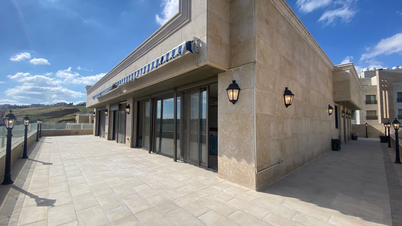 48fd9f94-320e-4632-8ff9-6bcf338f0cf7 Beige stone building with large glass doors on a sunlit terrace, striped blue-and-white awning, and black lantern lights along the wall.