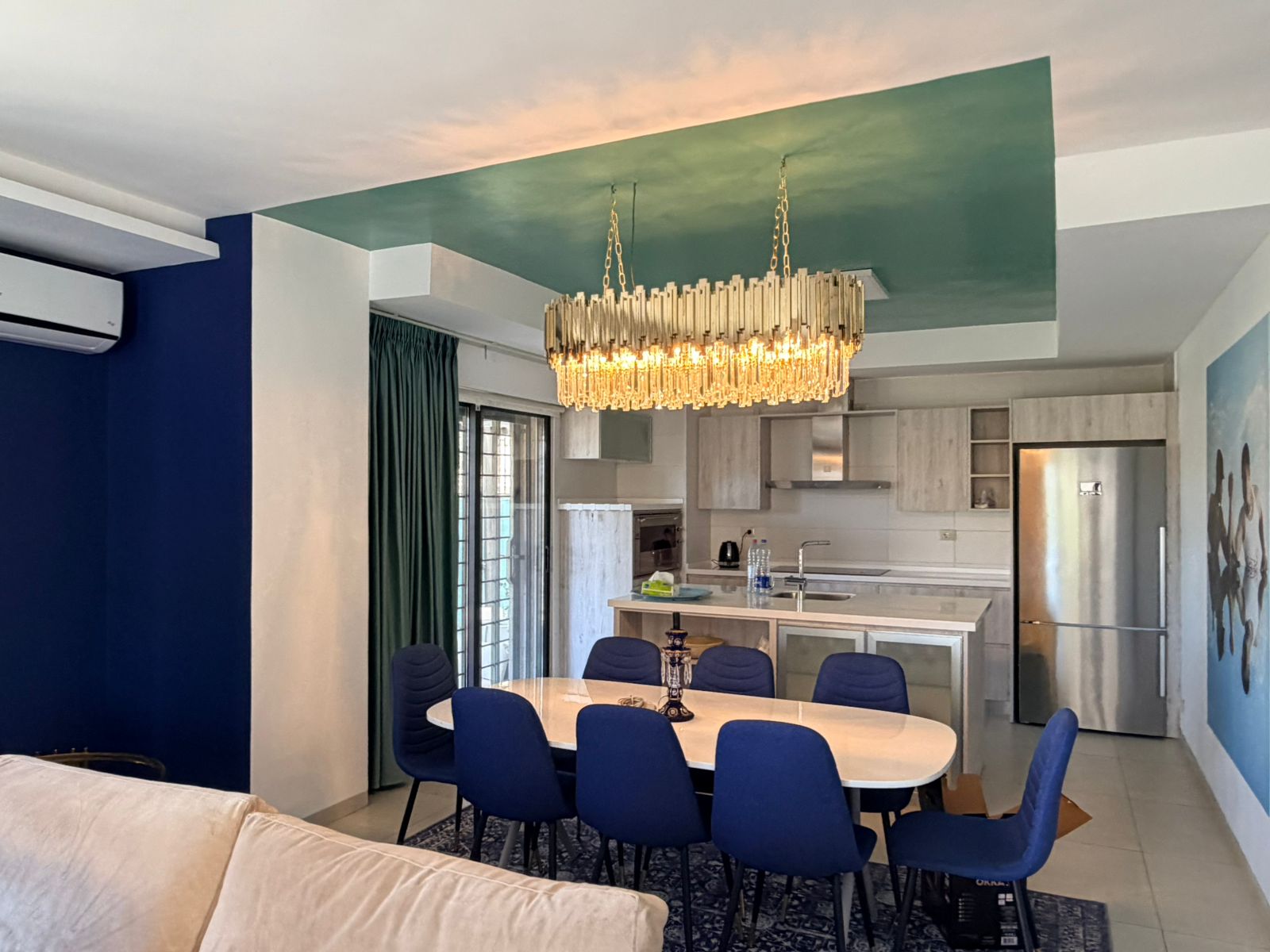 5009b856-9dd5-4afd-9bf6-5c65ed5aca72 Open-plan dining and kitchen area with a large rectangular white table surrounded by blue chairs and a crystal chandelier above the island.